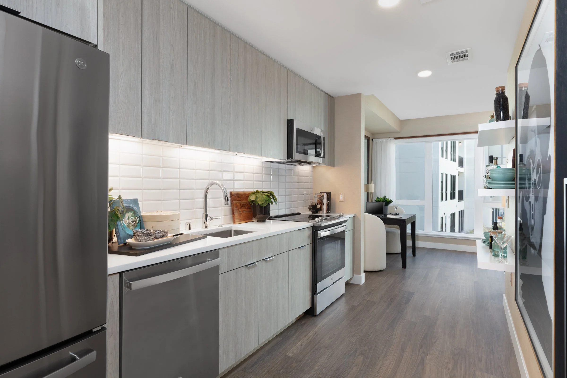 Modern kitchen with light wood cabinets, white subway tile backsplash, stainless steel refrigerator and appliances, black accents, and a view to a living area with large windows.