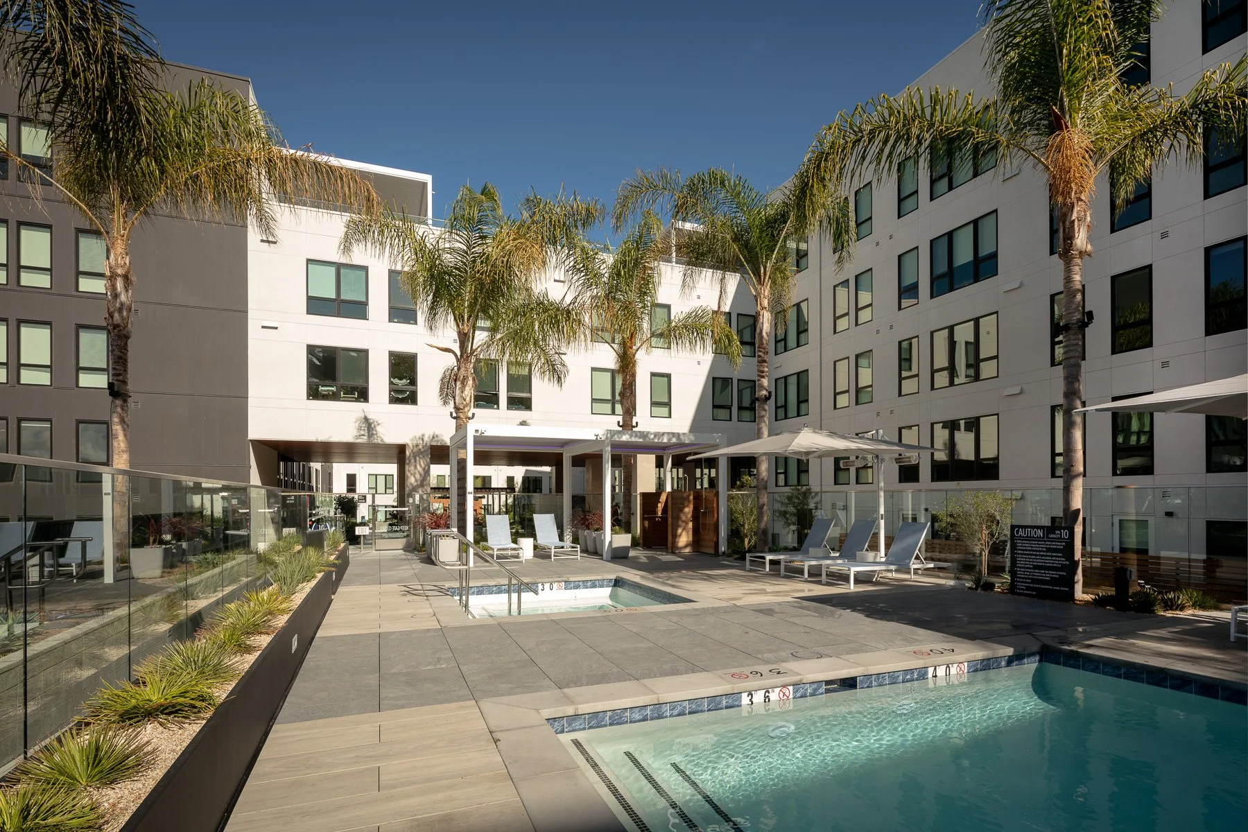 Outdoor pool area with lounge chairs, large white hotel building, palm trees, and umbrellas on a sunny day
