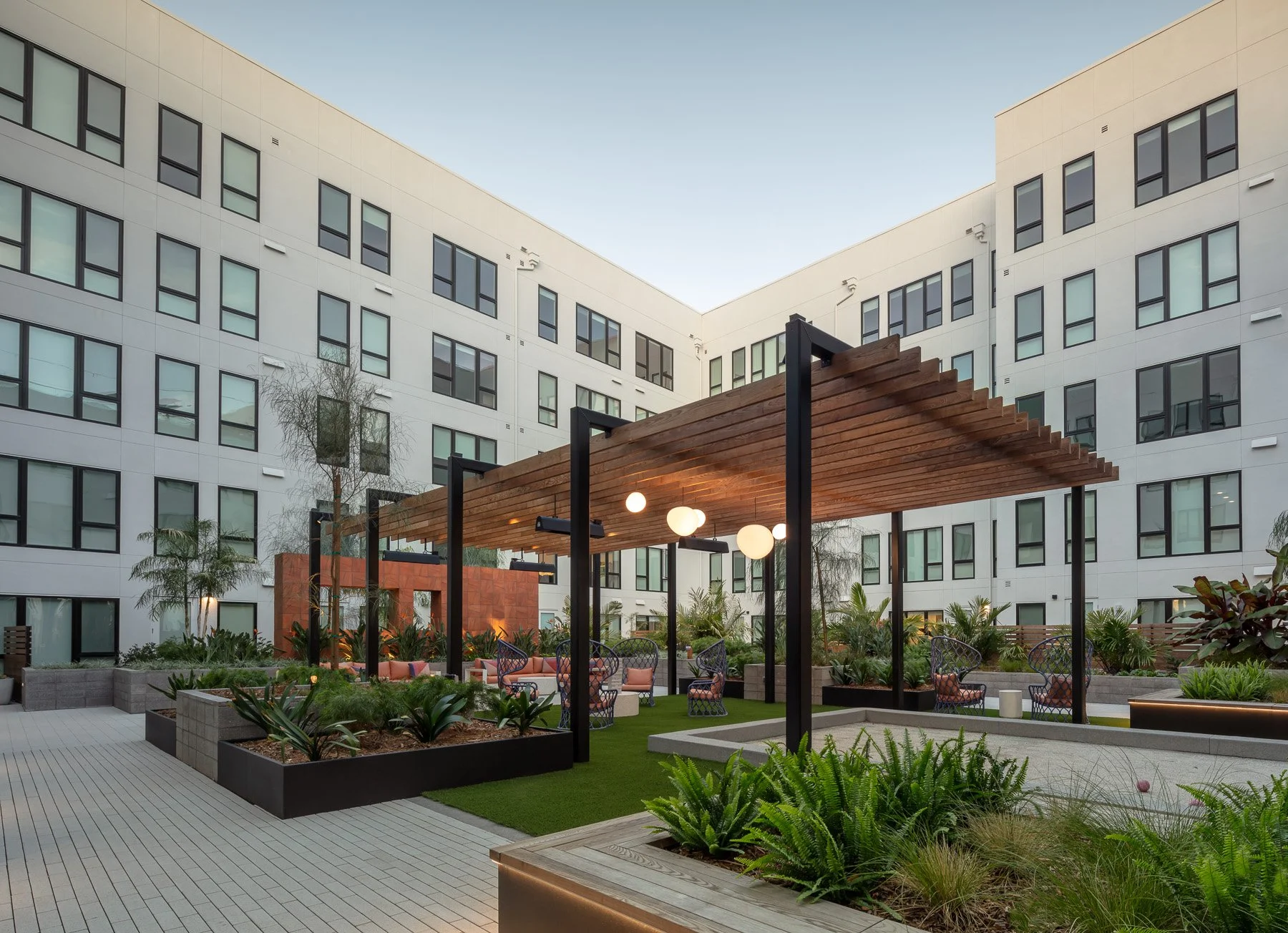 Outdoor courtyard with seating, garden beds, and a wooden pergola with hanging light fixtures, surrounded by modern white apartment buildings.