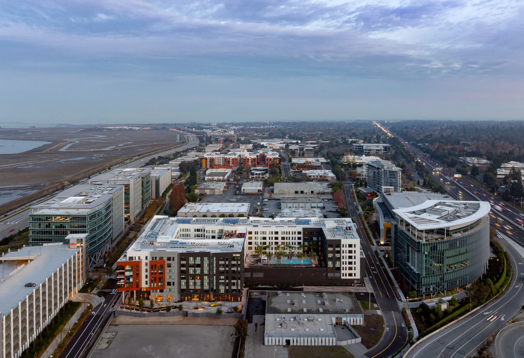Aerial view of a modern cityscape at dusk, featuring high-rise buildings, a highway with moving traffic, and open land or wetlands in the background.