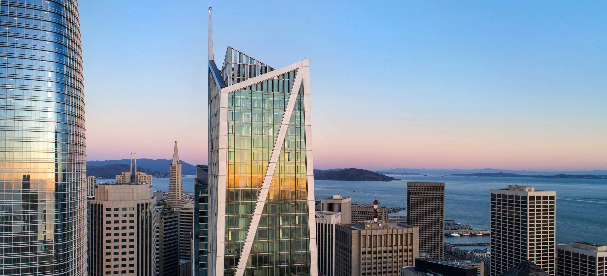 City skyline featuring tall modern glass buildings overlooking a bay at sunset, with mountains in the background.