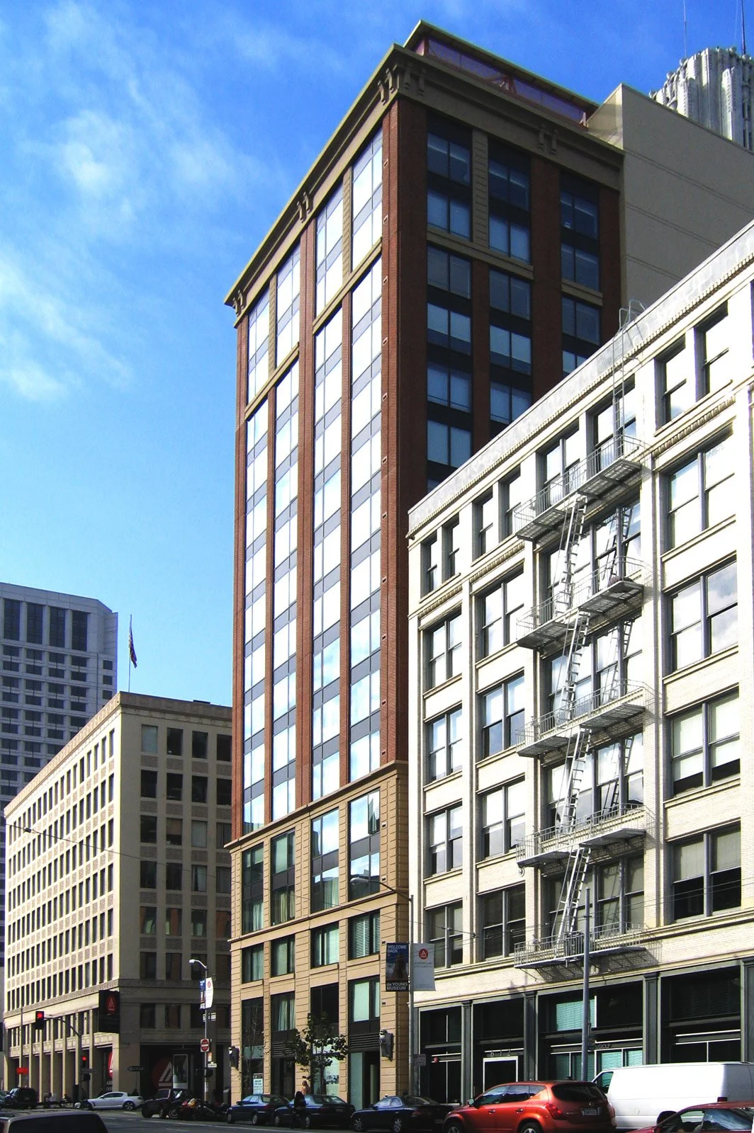 A city street with tall buildings, including a brown and glass skyscraper, a beige building with fire escape stairs, and low-rise shops with cars parked on the street.