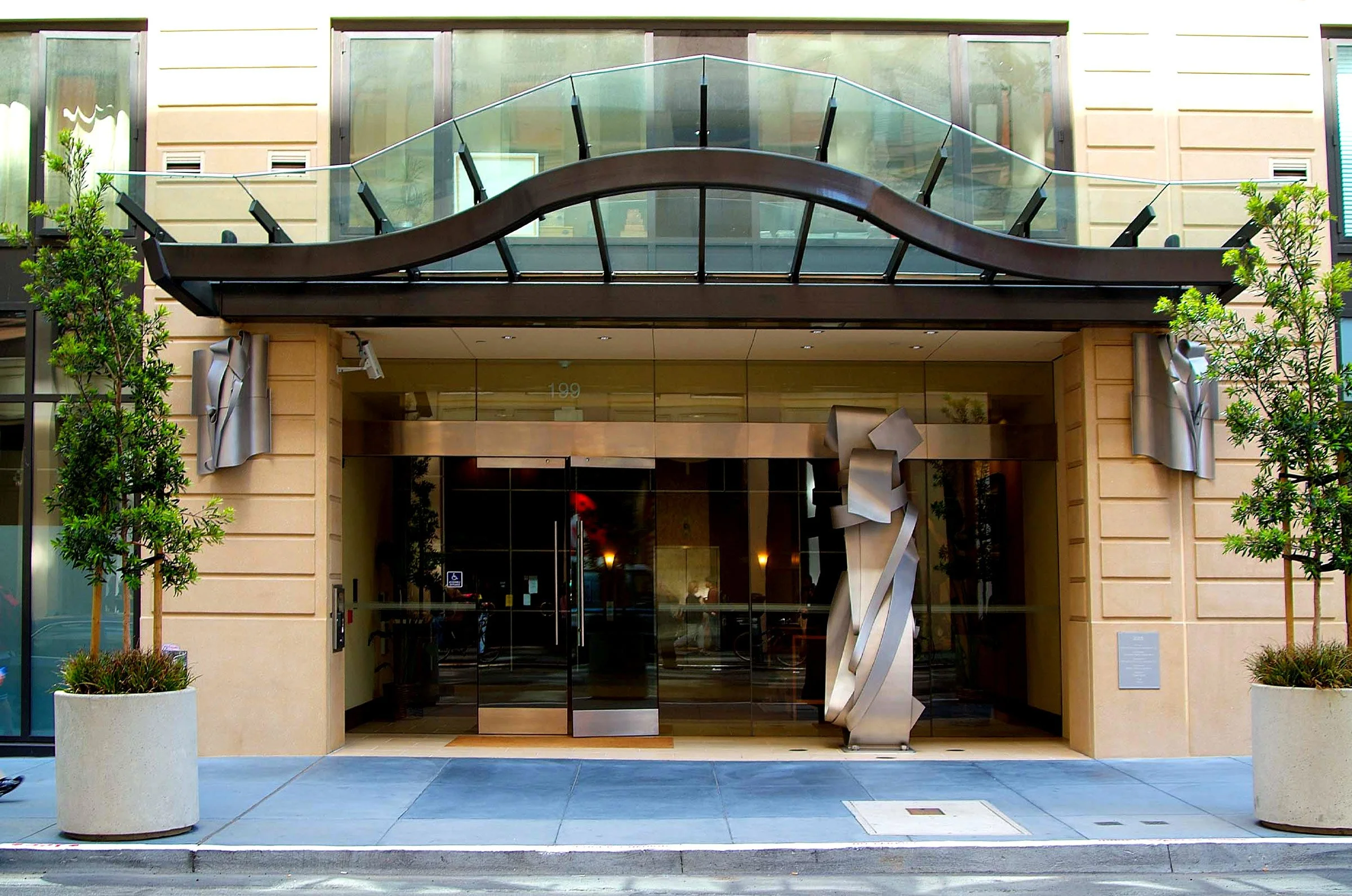 Modern building entrance with glass doors, metal sculptures, potted trees, and a glass balcony with a wooden wavy canopy.