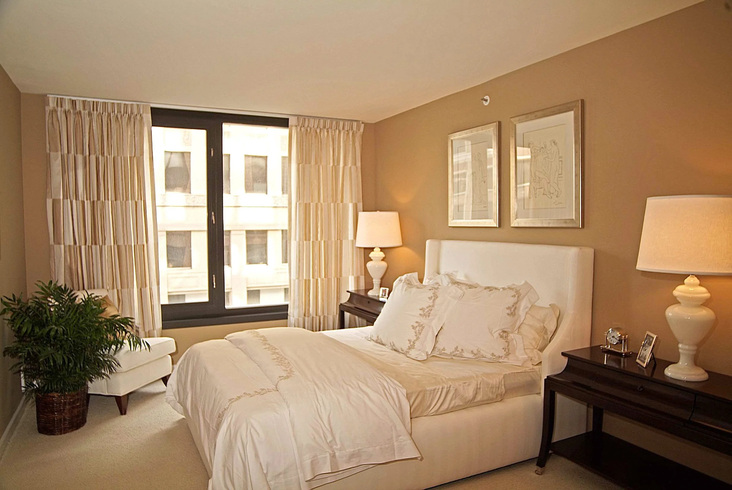 A neatly made bed with white bedding and decorative pillows in a cozy bedroom. There are two nightstands with lamps, framed artwork above the bed, a window with curtains, and a potted plant in the corner.
