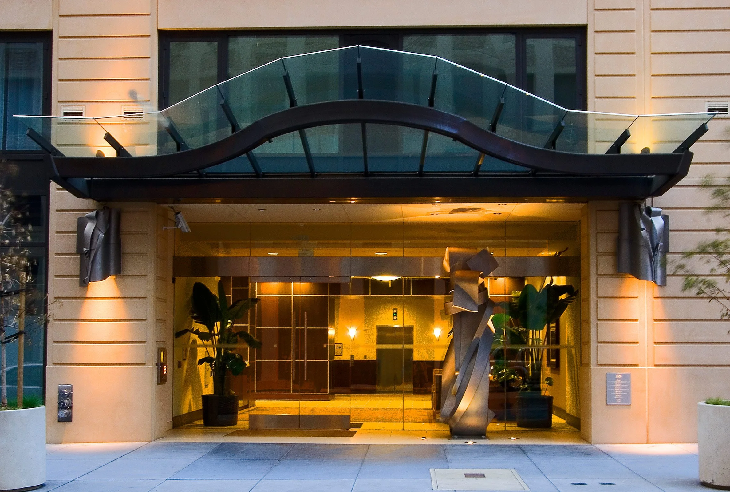 Entrance of a modern building with glass doors, potted plants on either side, and a decorative wave-shaped glass awning on top.