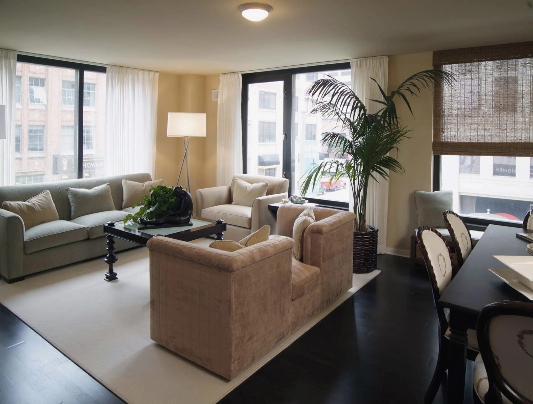 A modern living room with large windows, beige sofas, armchairs, a glass coffee table, a tall floor lamp, indoor plants, and a dining table with chairs.