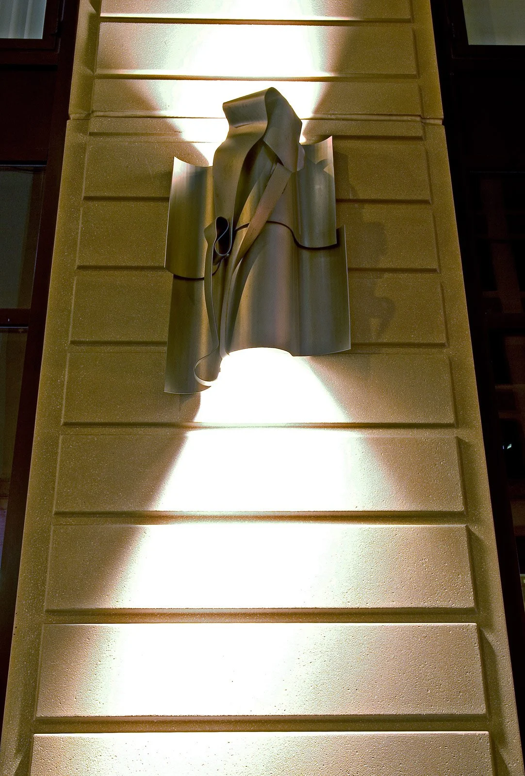 A metallic sculpture of a woman wearing a coat and holding a bag, mounted on a yellow textured wall with lighting creating shadows.