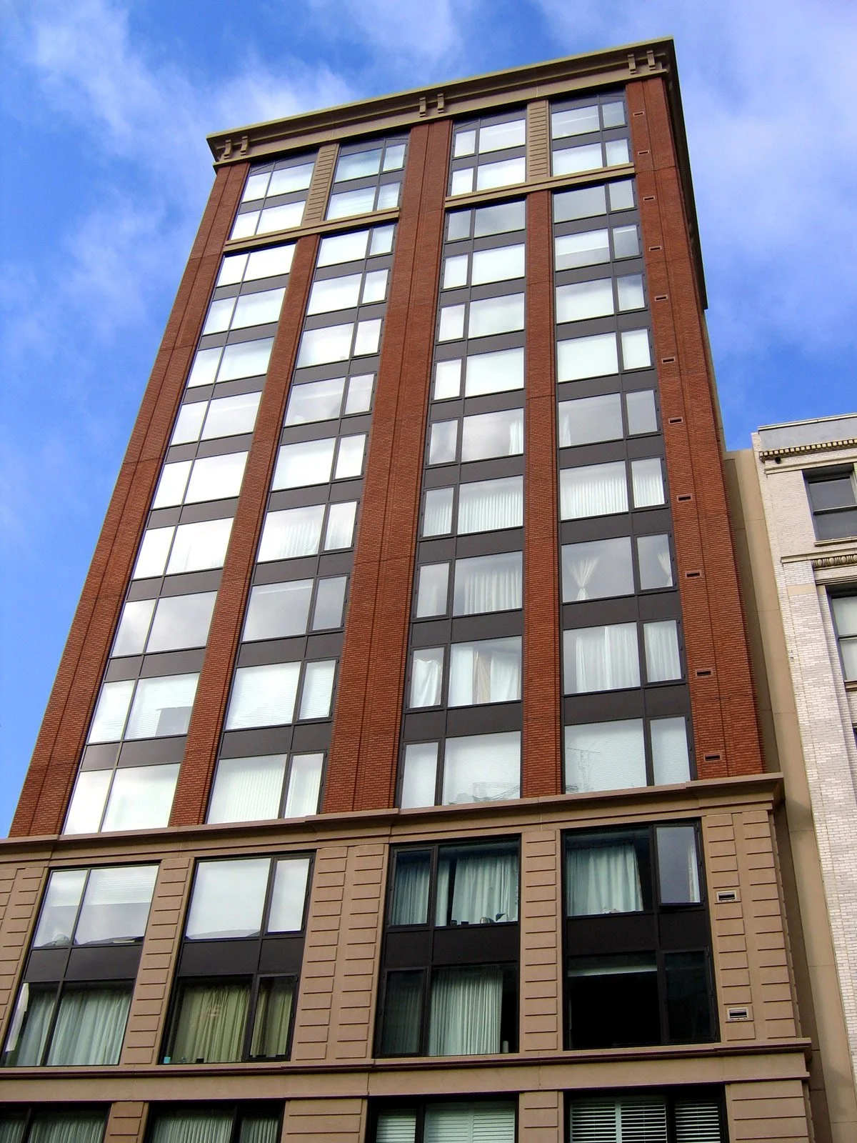 A tall multi-story building with a brick and glass facade, featuring large windows and vertical design elements, against a partly cloudy sky.