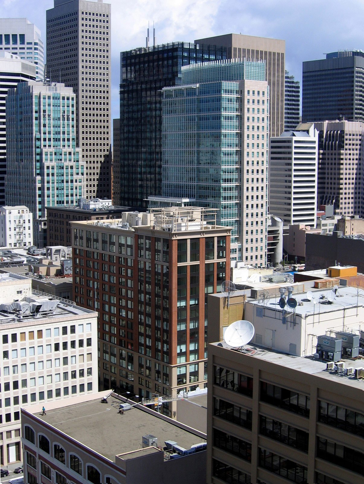 A cityscape with tall modern skyscrapers and buildings, featuring glass facades and rooftop satellite dishes, under a partly cloudy sky.