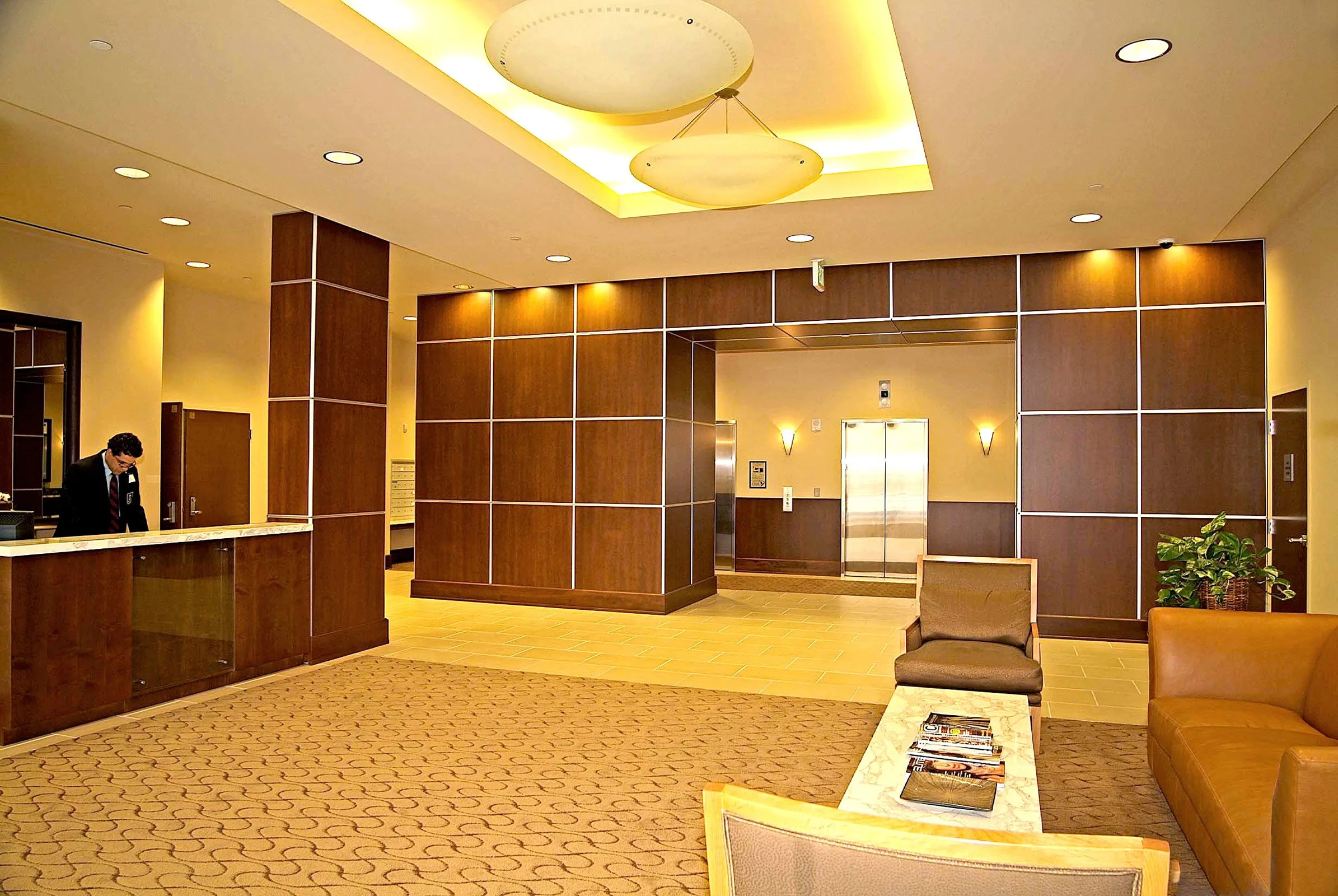 Hotel lobby with reception desk, seating area with chairs and coffee table, elevator, and warm lighting.