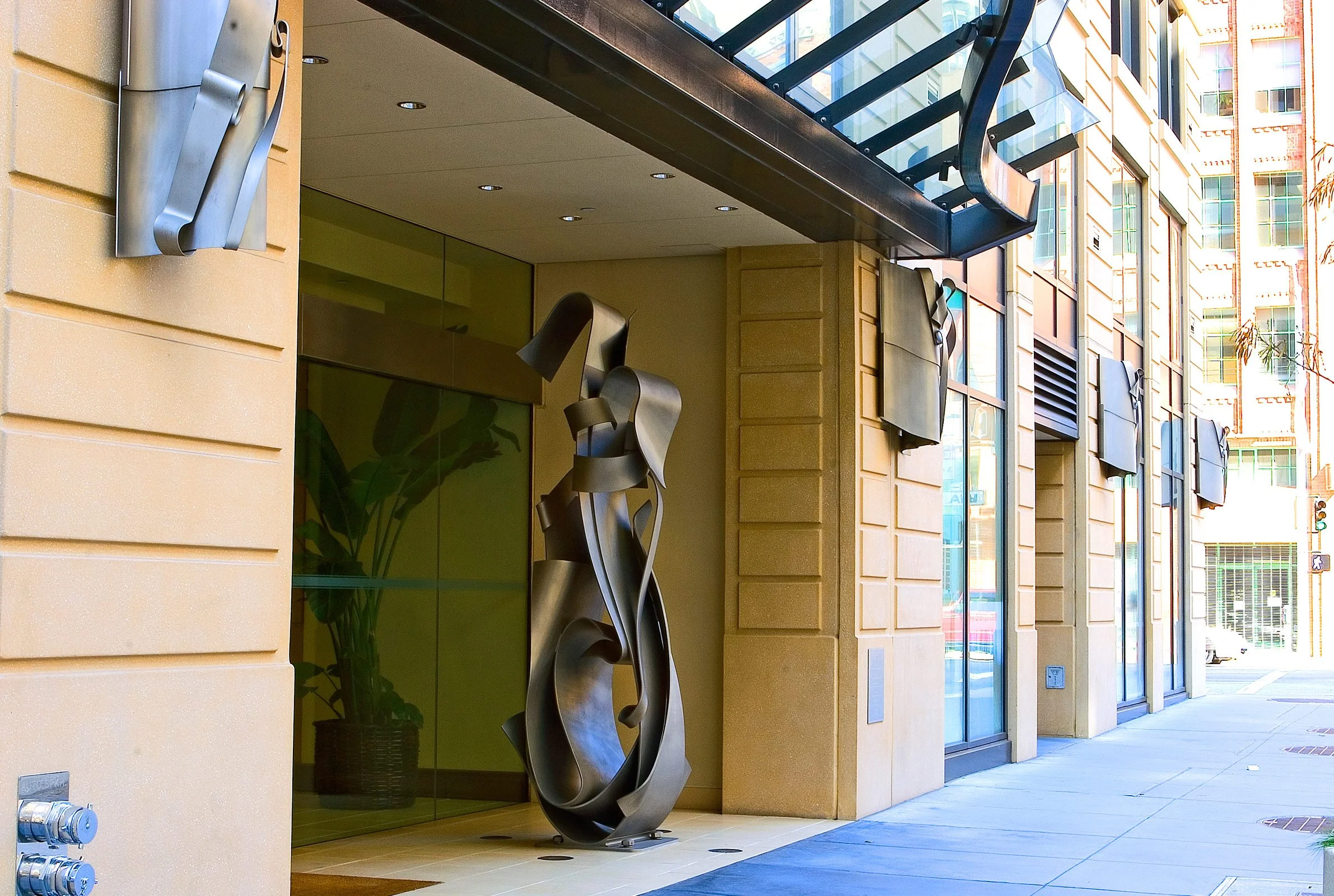 Modern building entrance with abstract metal sculpture and glass door, beige walls, and large windows reflecting city surroundings.