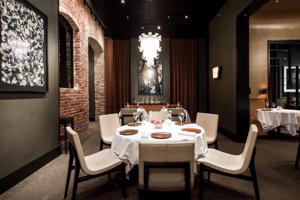 Elegant restaurant dining room with brick walls, modern artwork, white tablecloths, and a chandelier.