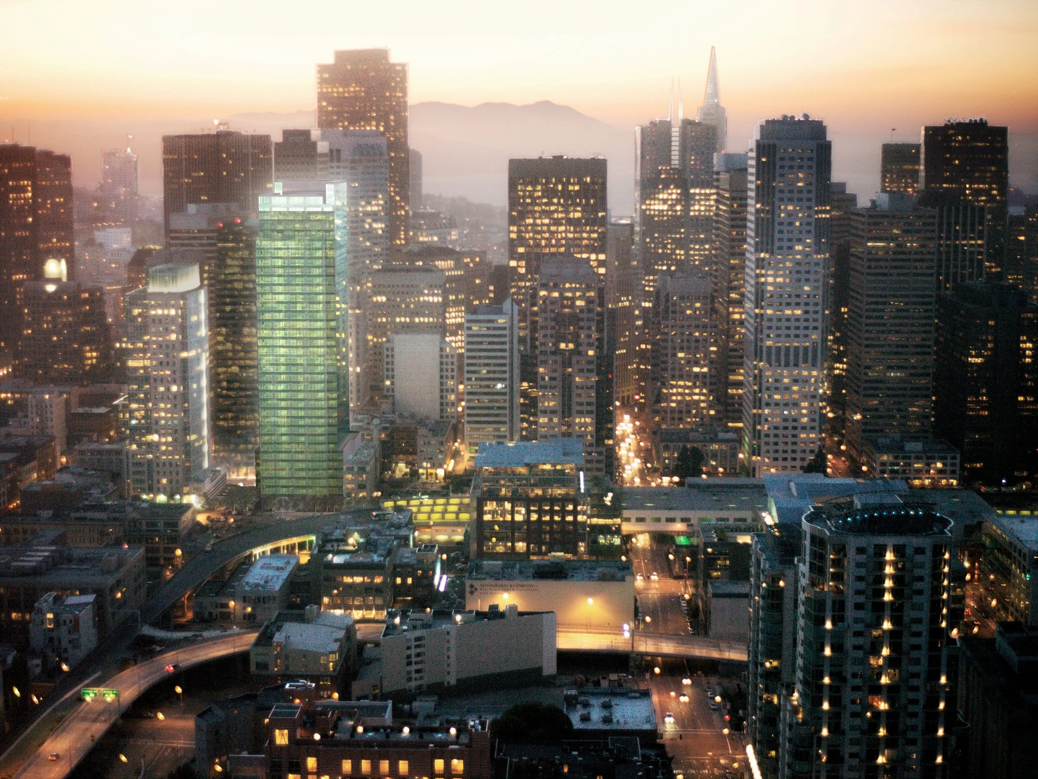 Cityscape of downtown San Francisco at dusk with illuminated skyscrapers, including the Transamerica Pyramid, against a background of mountains and a sky transitioning from sunset to night.