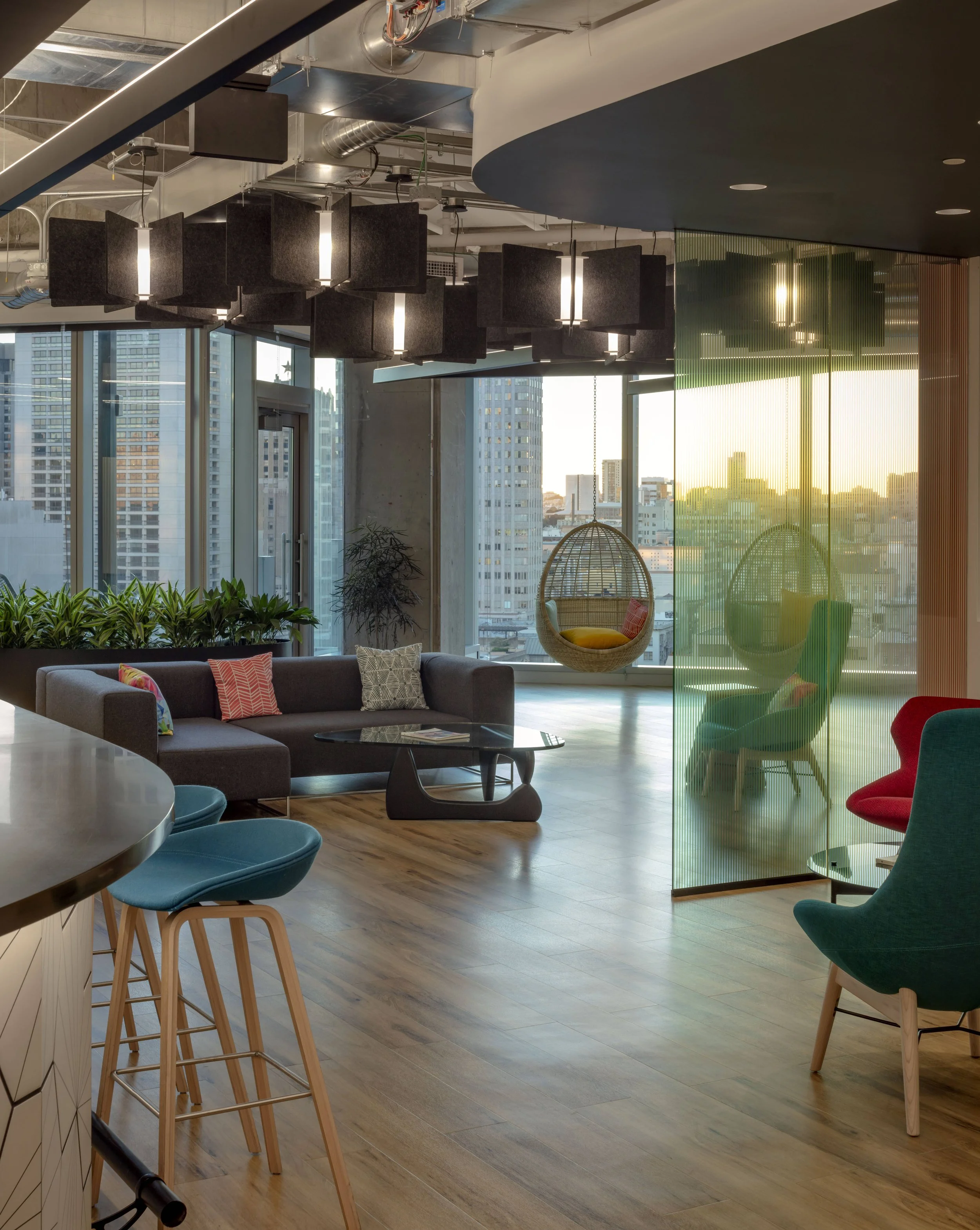 Modern city view lounge with colorful chairs, a hanging egg chair, a sofa with decorative pillows, and large windows showing cityscape at sunset.