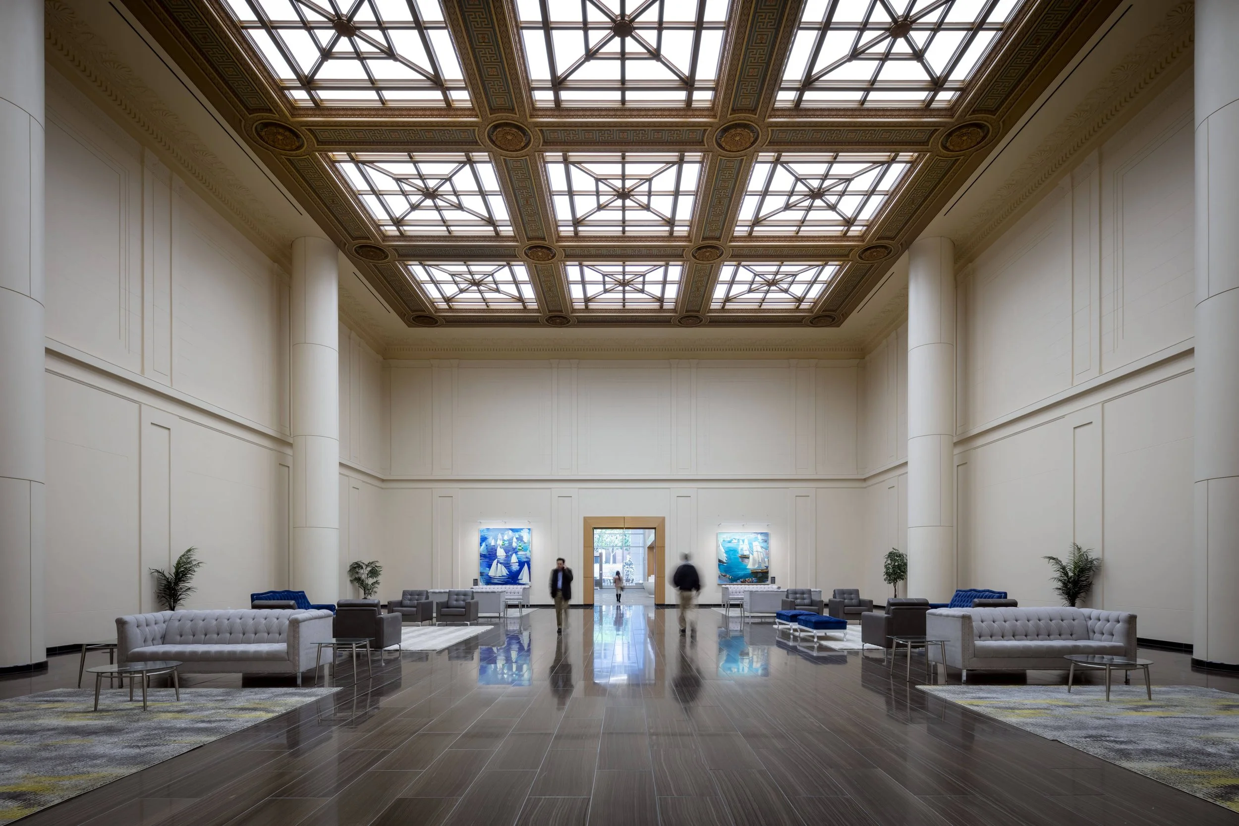 A spacious, elegant lobby with high ceiling featuring decorative skylights, white walls, and dark wood flooring. There are several sofas and chairs arranged around small tables, with potted plants on either side. Artworks are displayed on the walls, 