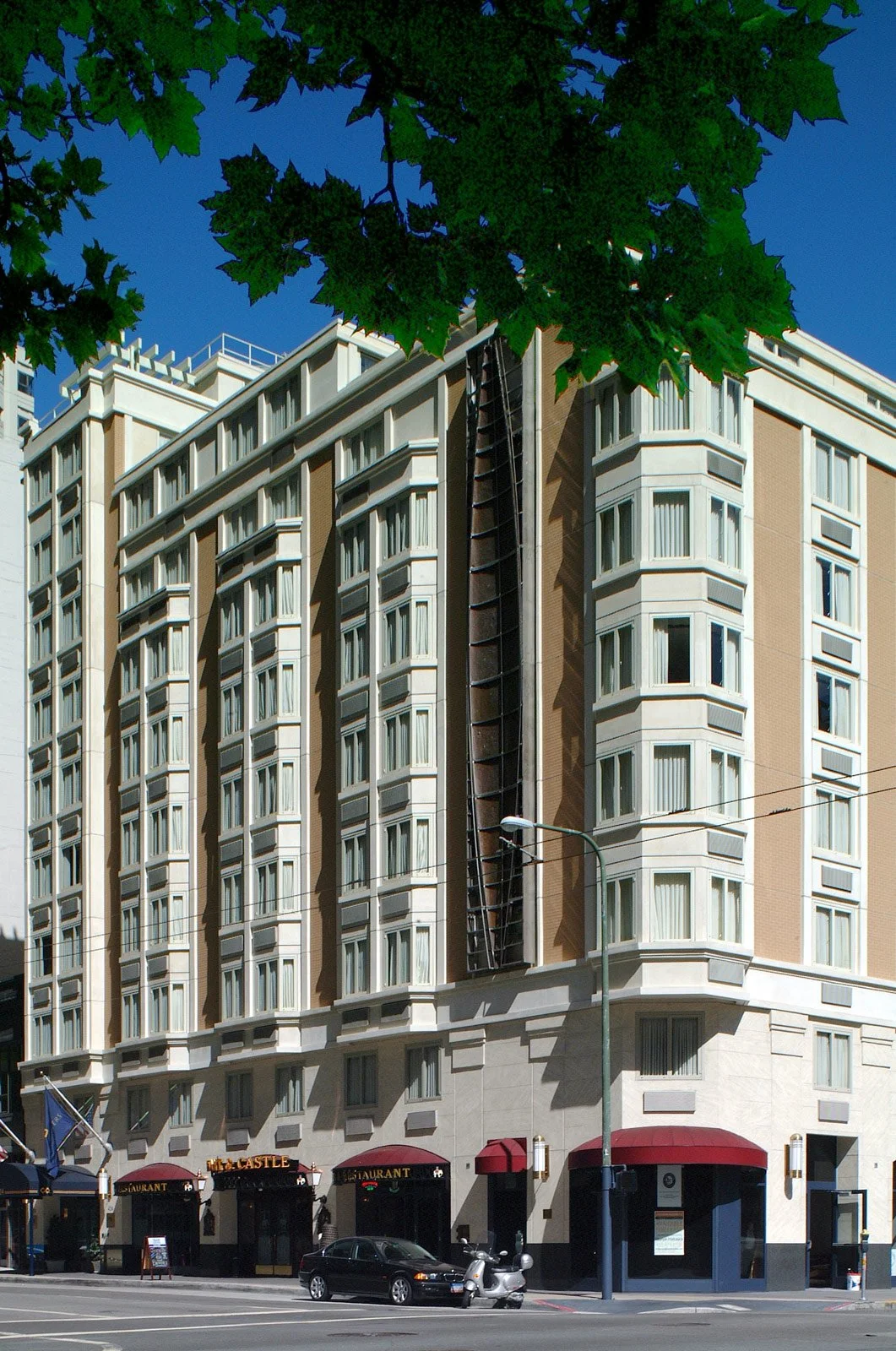 A multi-story building with a restaurant at the ground level, parked cars, a street lamp, and green tree leaves in the foreground. The building has numerous windows and a vertical black structure in the center.