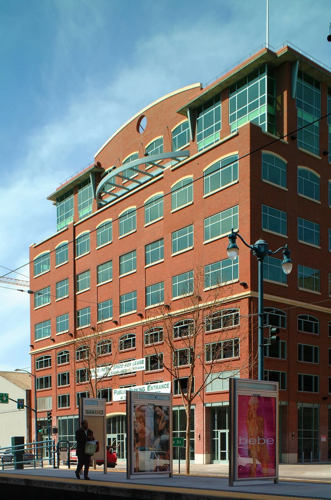 A multi-story red brick building with large windows and modern design elements above a city street, with a person at a bus stop, trees, street signs, and advertising posters.