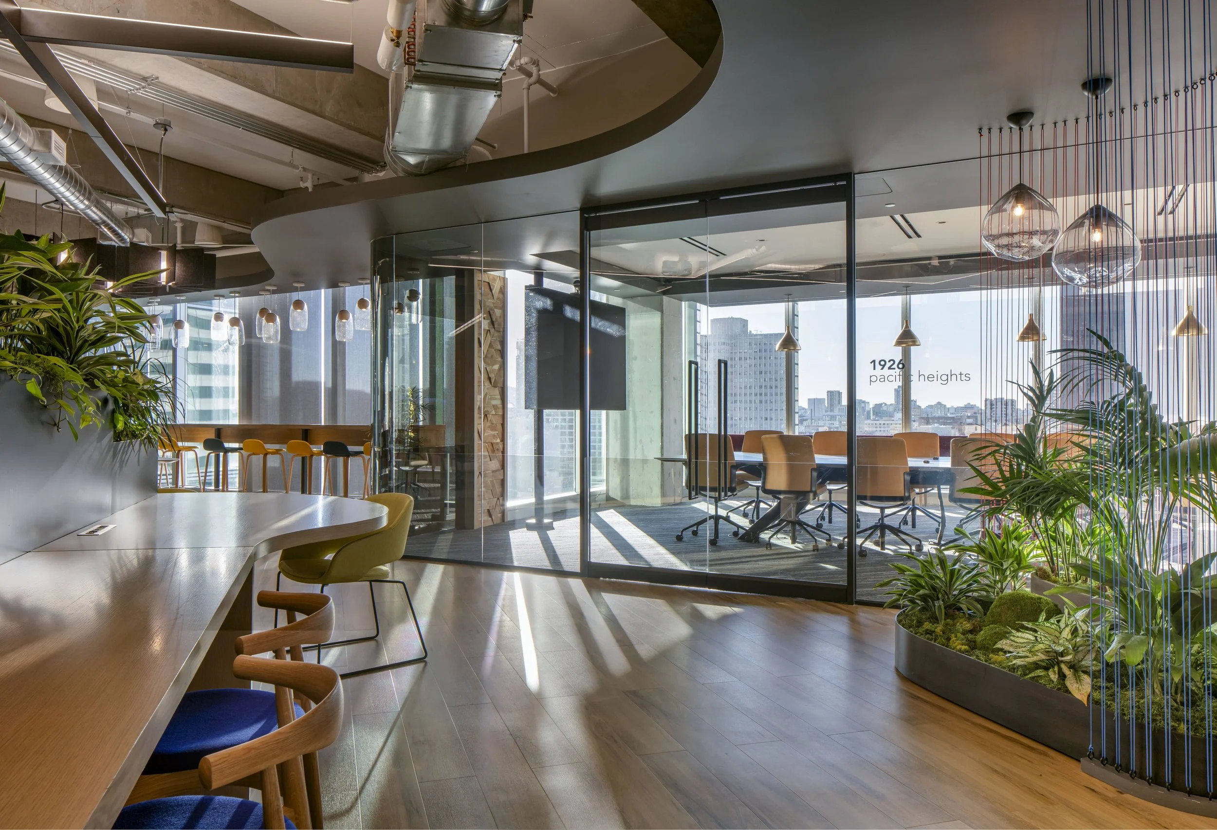 A modern office space with a glass conference room, wooden floors, and an adjacent area with a long table and colorful chairs. There are large windows letting in natural light, and there are plants and lighting fixtures hanging from the ceiling.