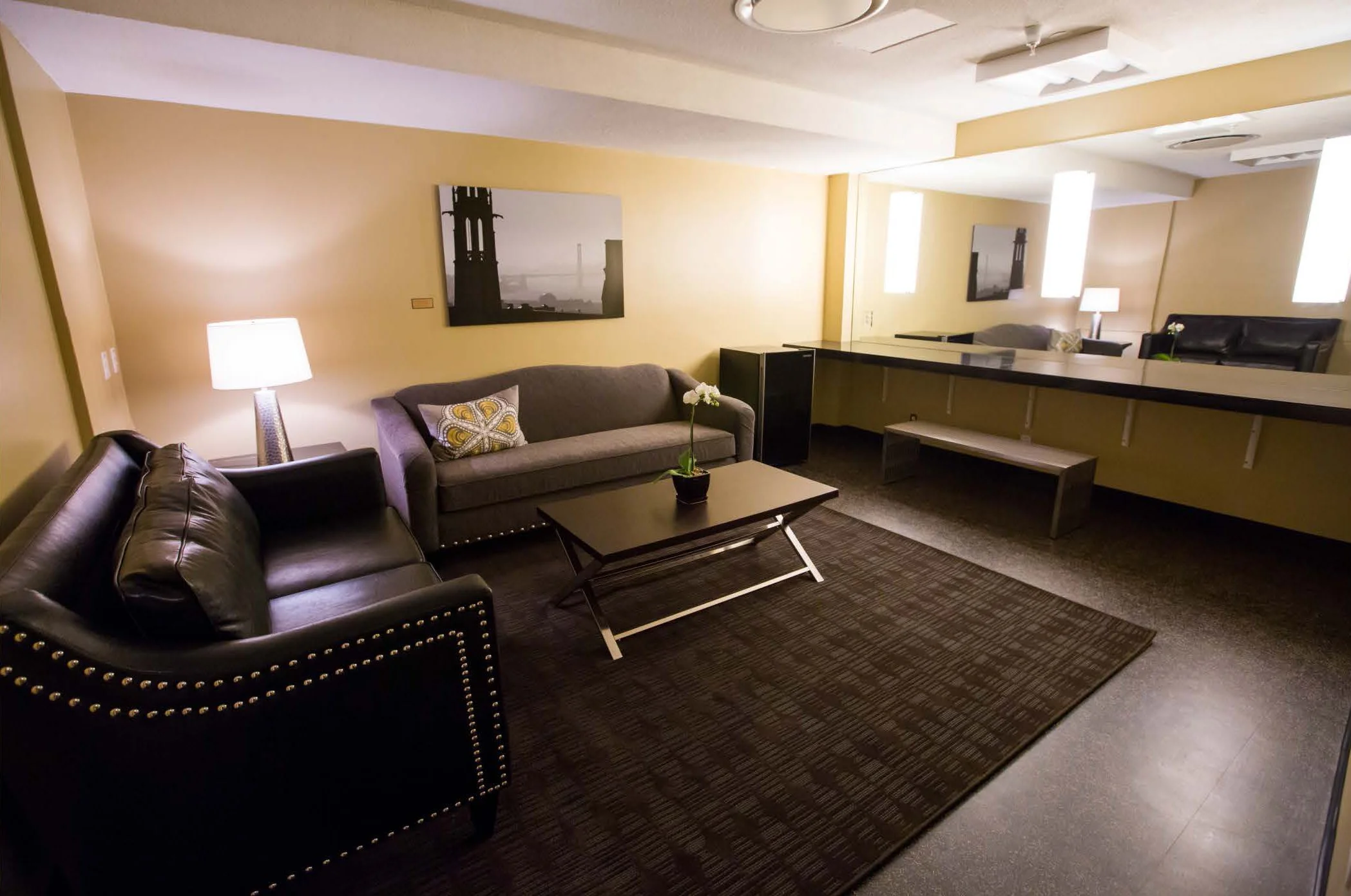 Cozy hotel lobby with brown leather and fabric sofas, a coffee table with a potted plant, lamps, and wall art, with warm lighting and carpeted flooring.