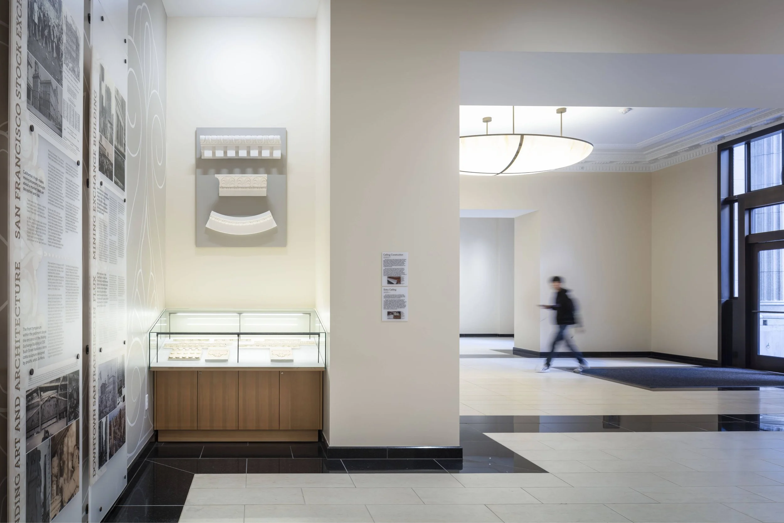 Interior of a museum with informational panels, a display case, a person walking and using a phone, and modern lighting fixtures.