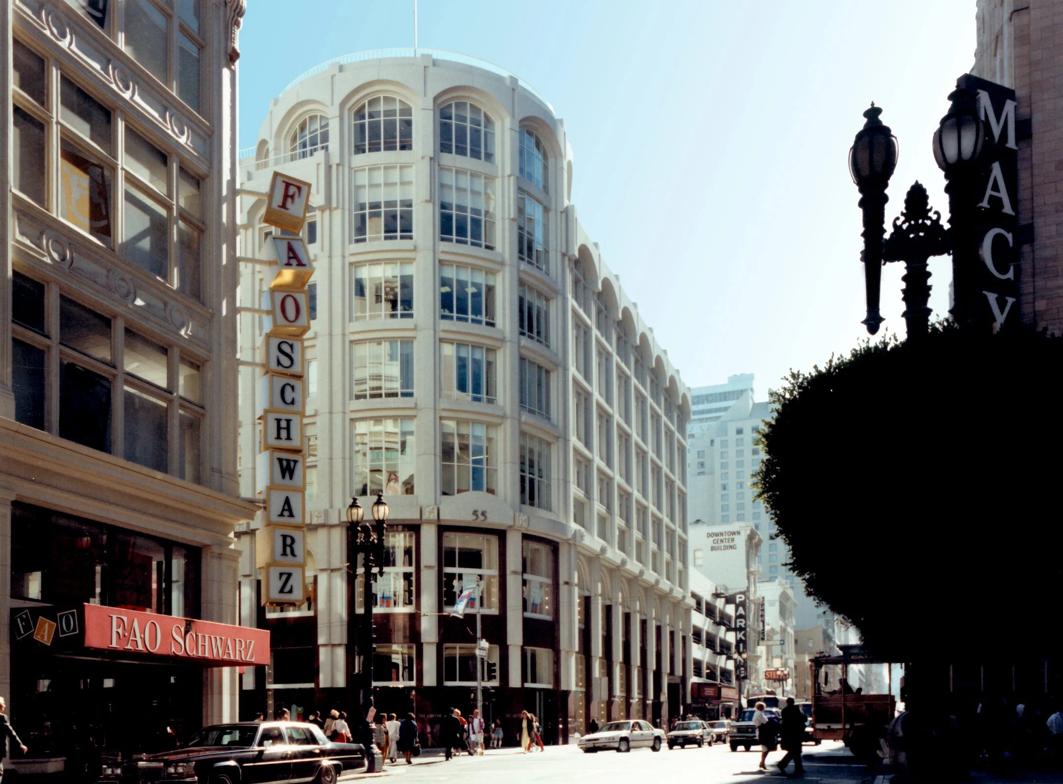 City street scene with tall buildings, including one with a sign reading FAO Schwarz, and people walking on the sidewalk with cars parked and driving by.