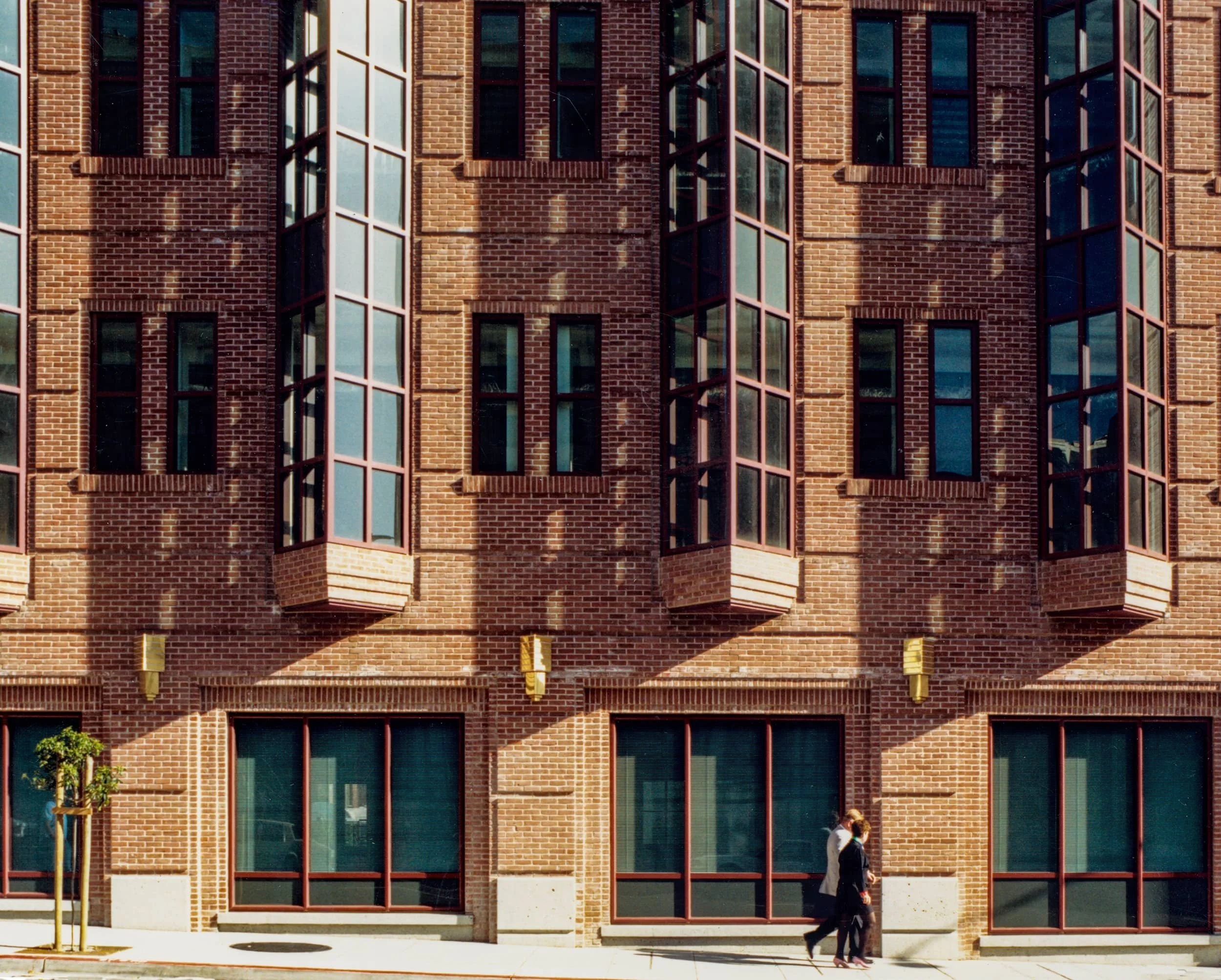 A brick building with large bay windows and reflective glass, with two people walking on the sidewalk in front of it.