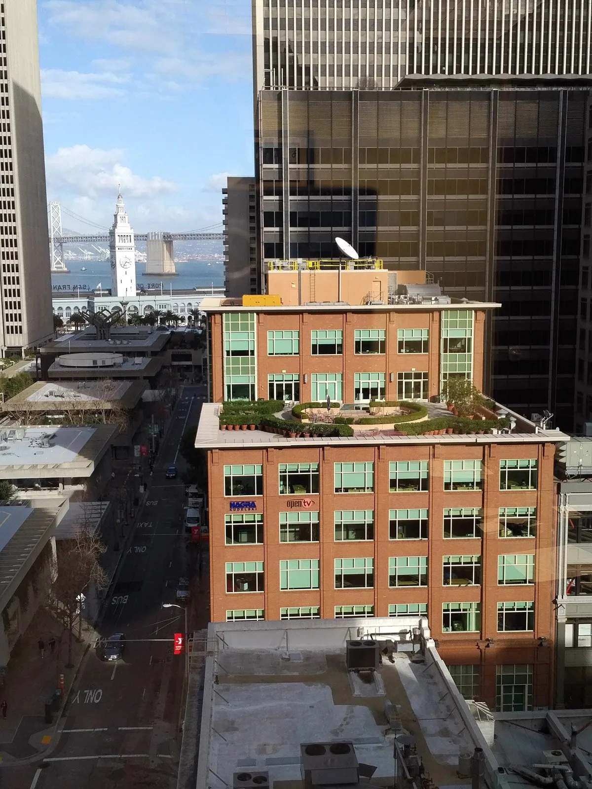 View of a cityscape with high-rise buildings, a rooftop garden, and the San Francisco Bay in the background with the Bay Bridge and a clock tower.