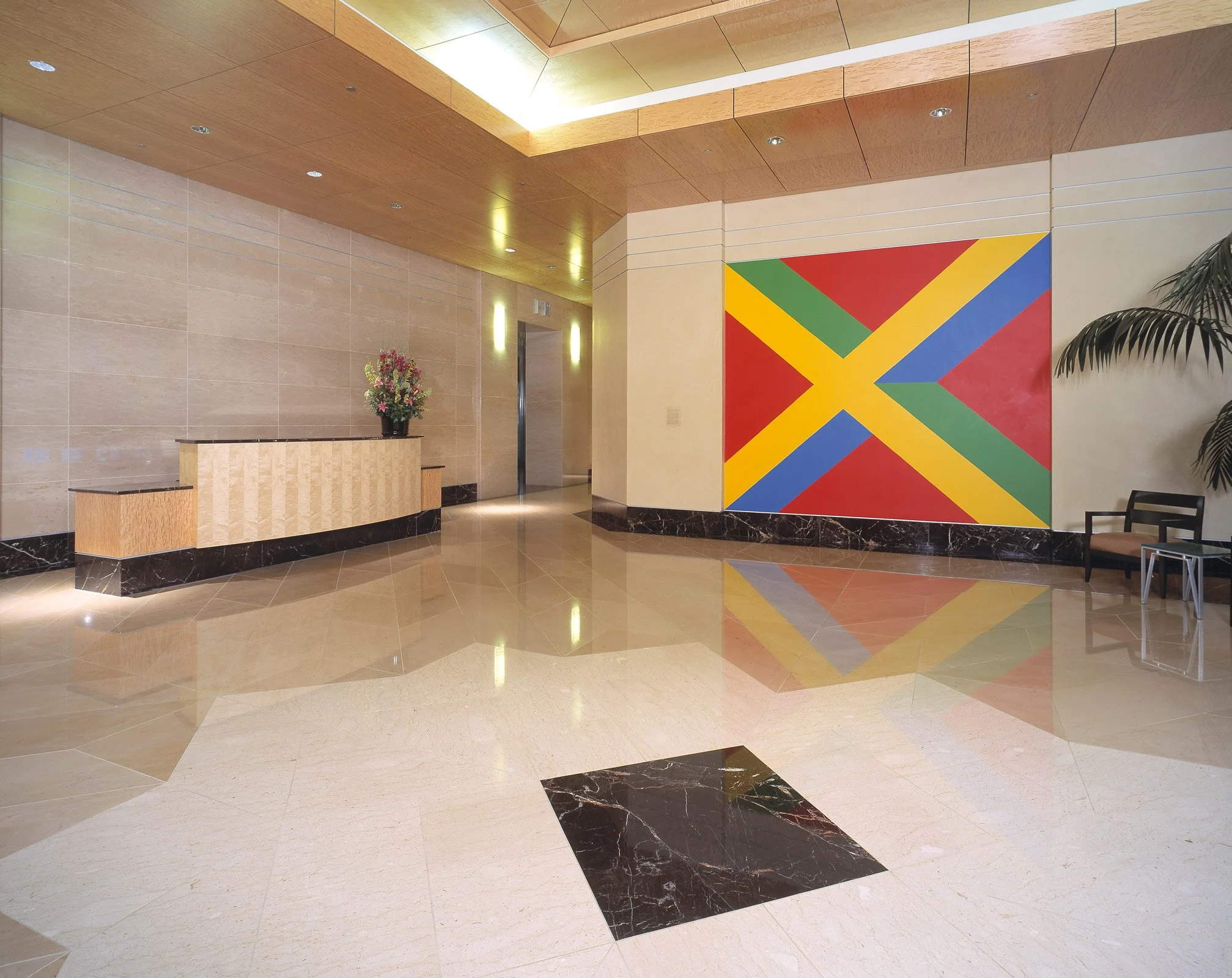 Hotel lobby with beige tiled walls and floors, a black marble border, a wooden reception desk with a flower arrangement, an elevator, and a large mural of the Jamaican flag on the wall.