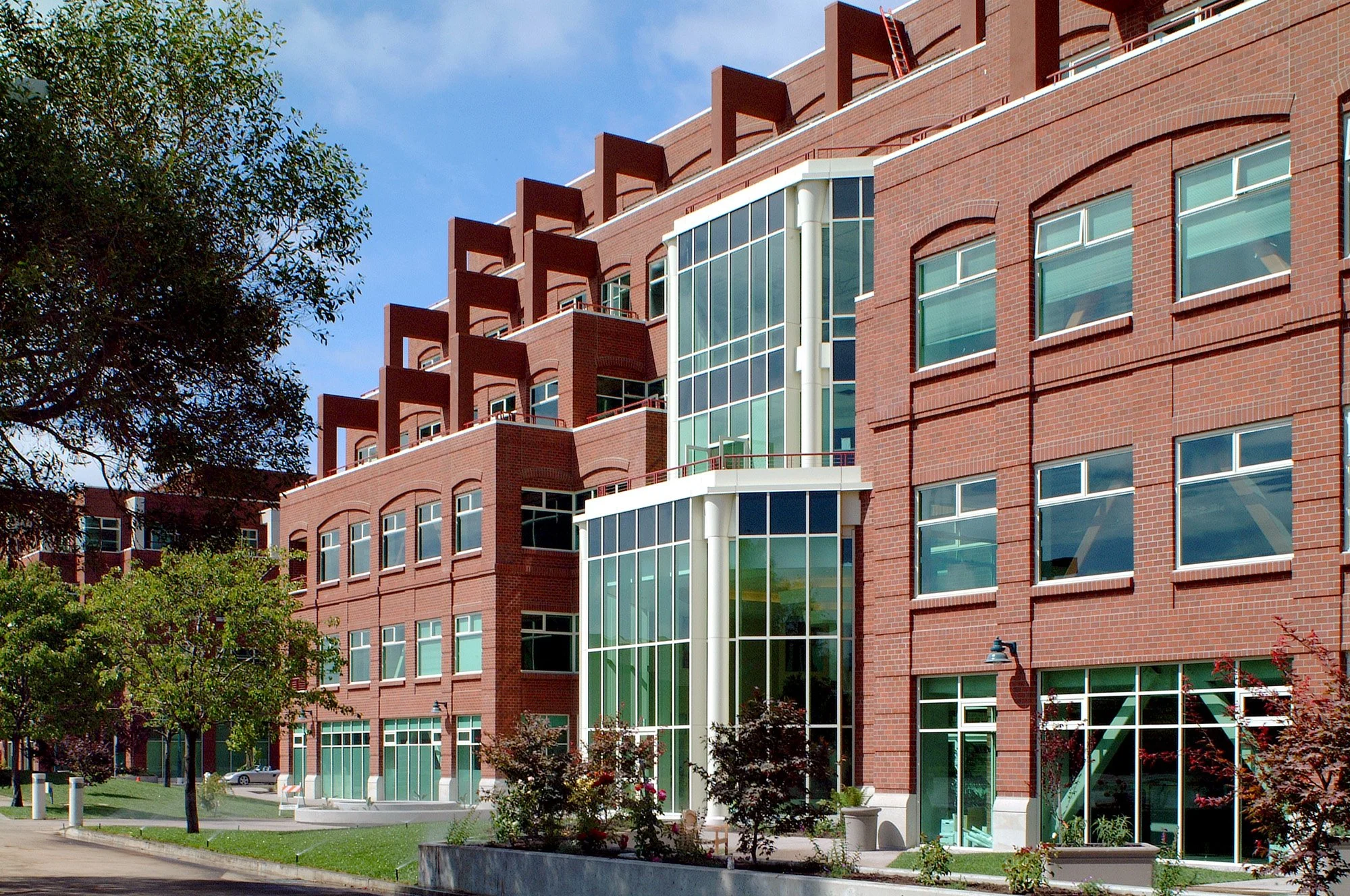 Modern multi-story building with red brick exterior and large glass windows, surrounded by trees and landscaped greenery.