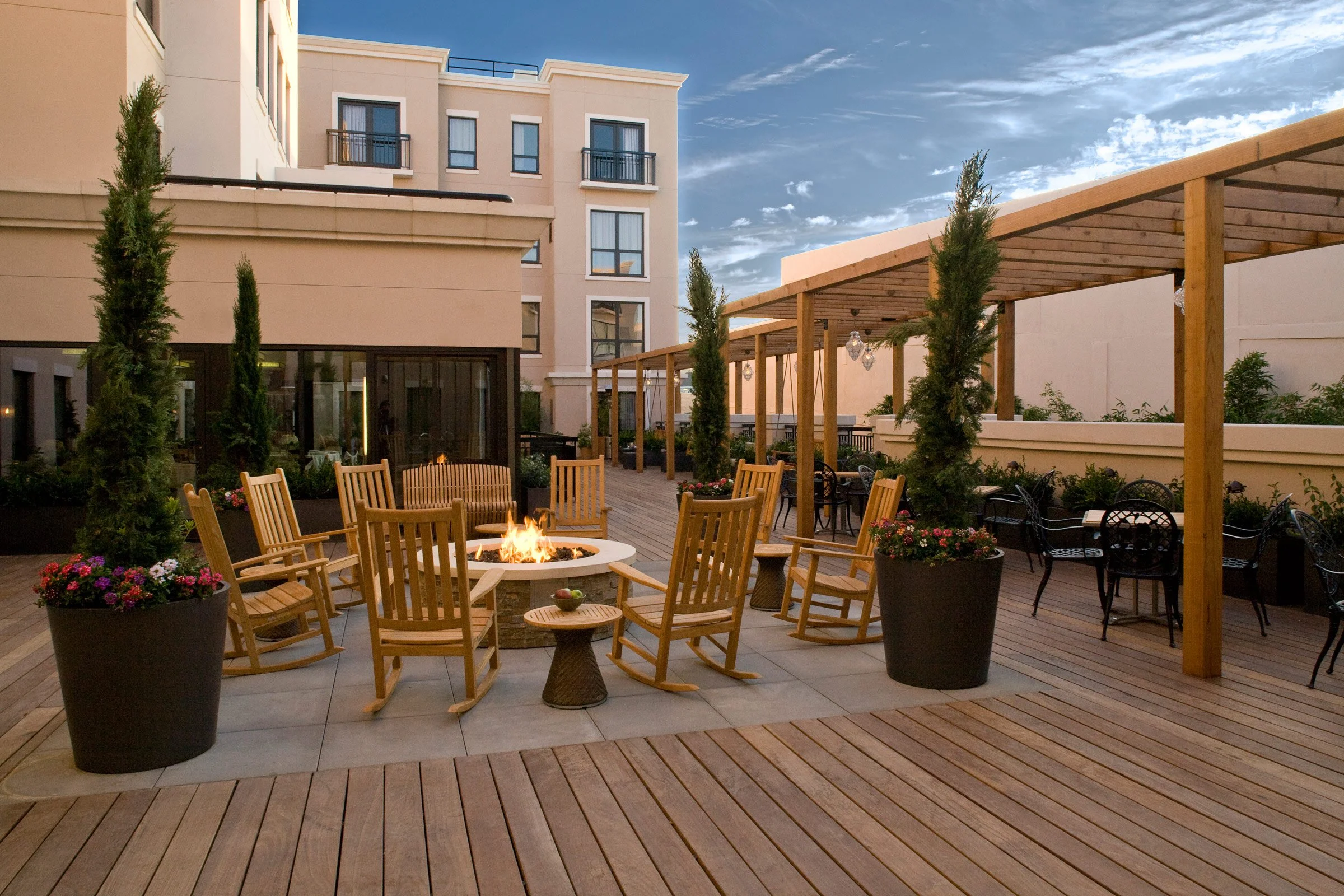 A rooftop patio with wooden decking, a fire pit surrounded by wooden chairs, decorated with potted plants, and a shaded seating area under a wooden pergola, with a multi-story building and a partly cloudy sky in the background.