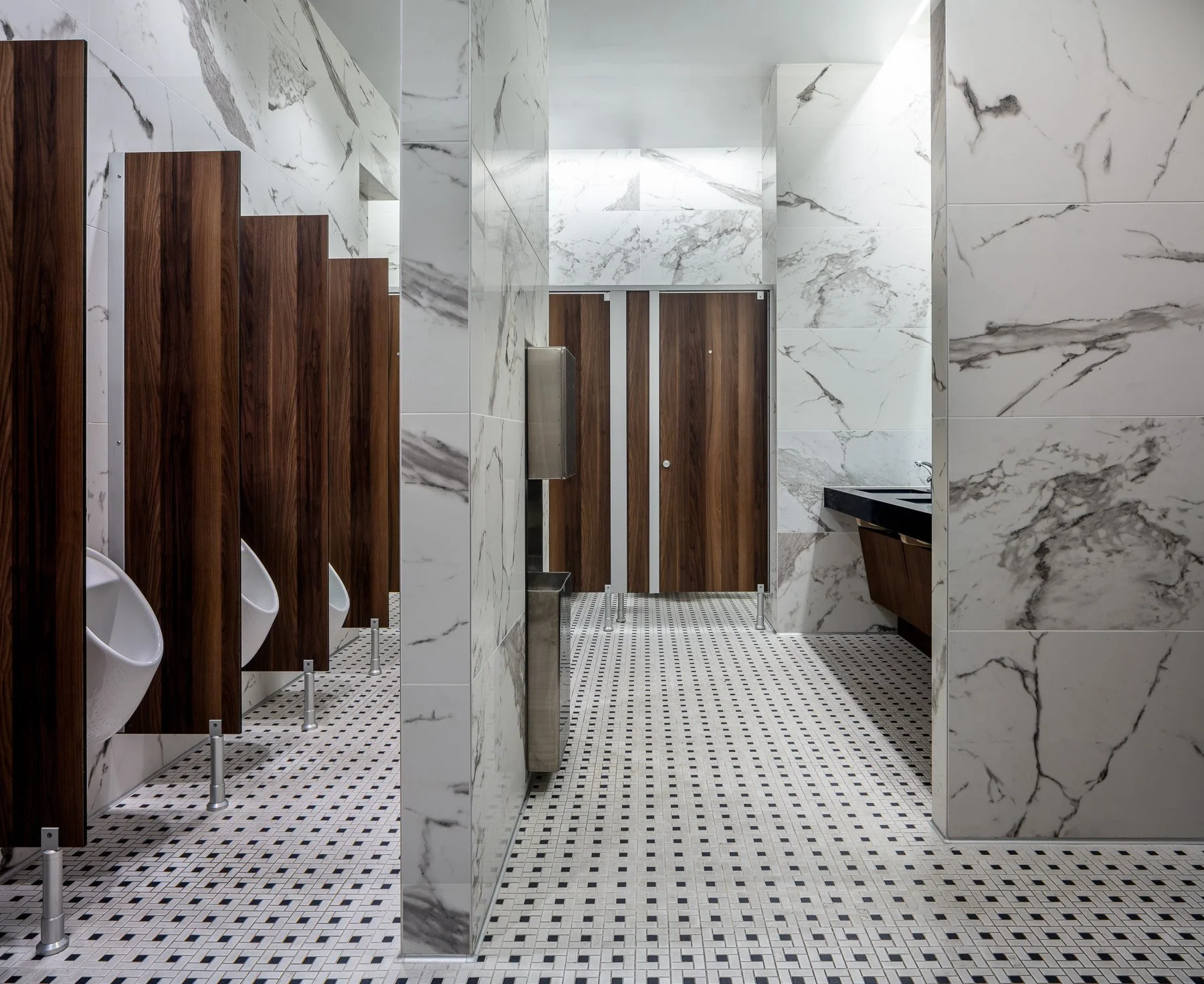 Public men's restroom with urinals, wooden stall partitions, and a sink with a black countertop. The walls are covered in white marble tiles with gray veining. The floor has small black and white mosaic tiles.