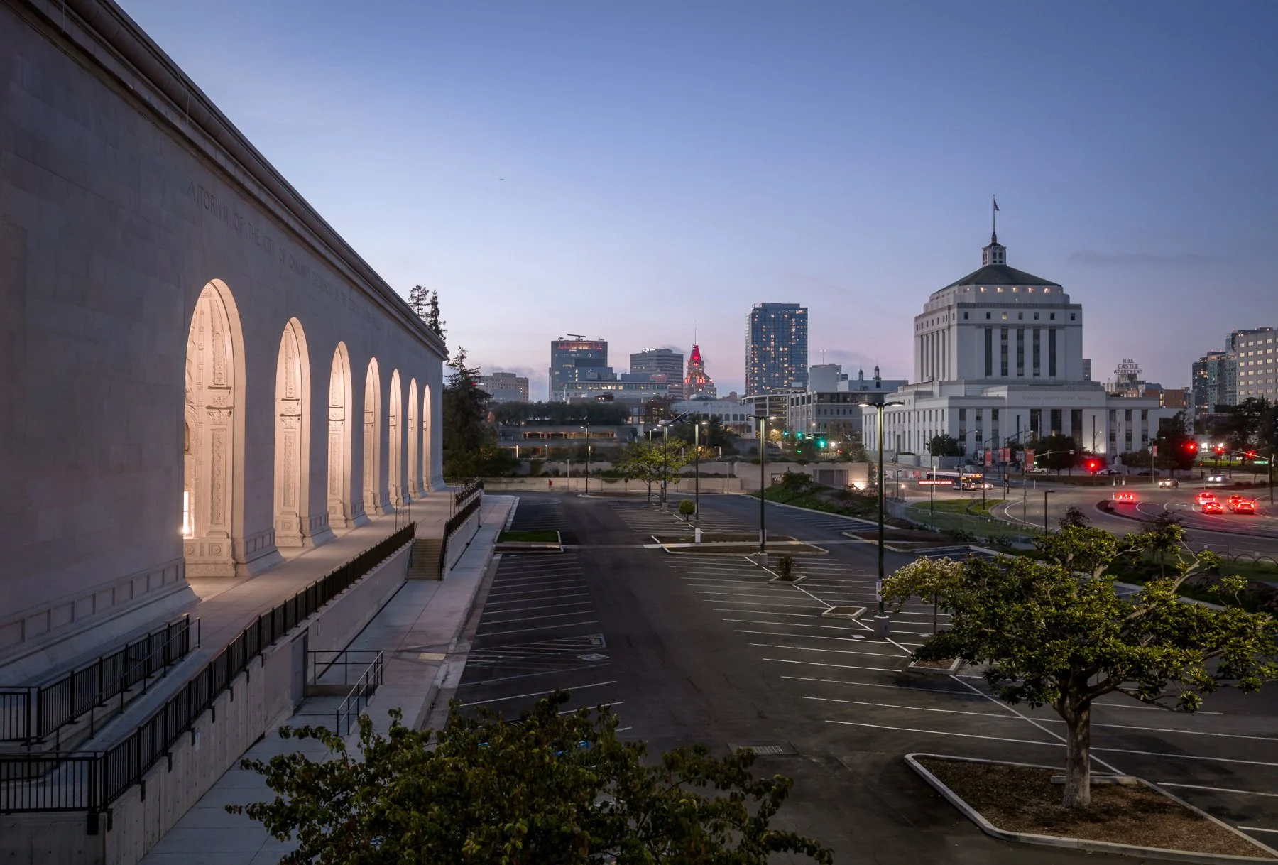 Cityscape at dusk with an empty parking lot in the foreground, a large white government building with columns on the right, and a skyline with various tall buildings in the background, including one with red lighting and a building with a flag on top