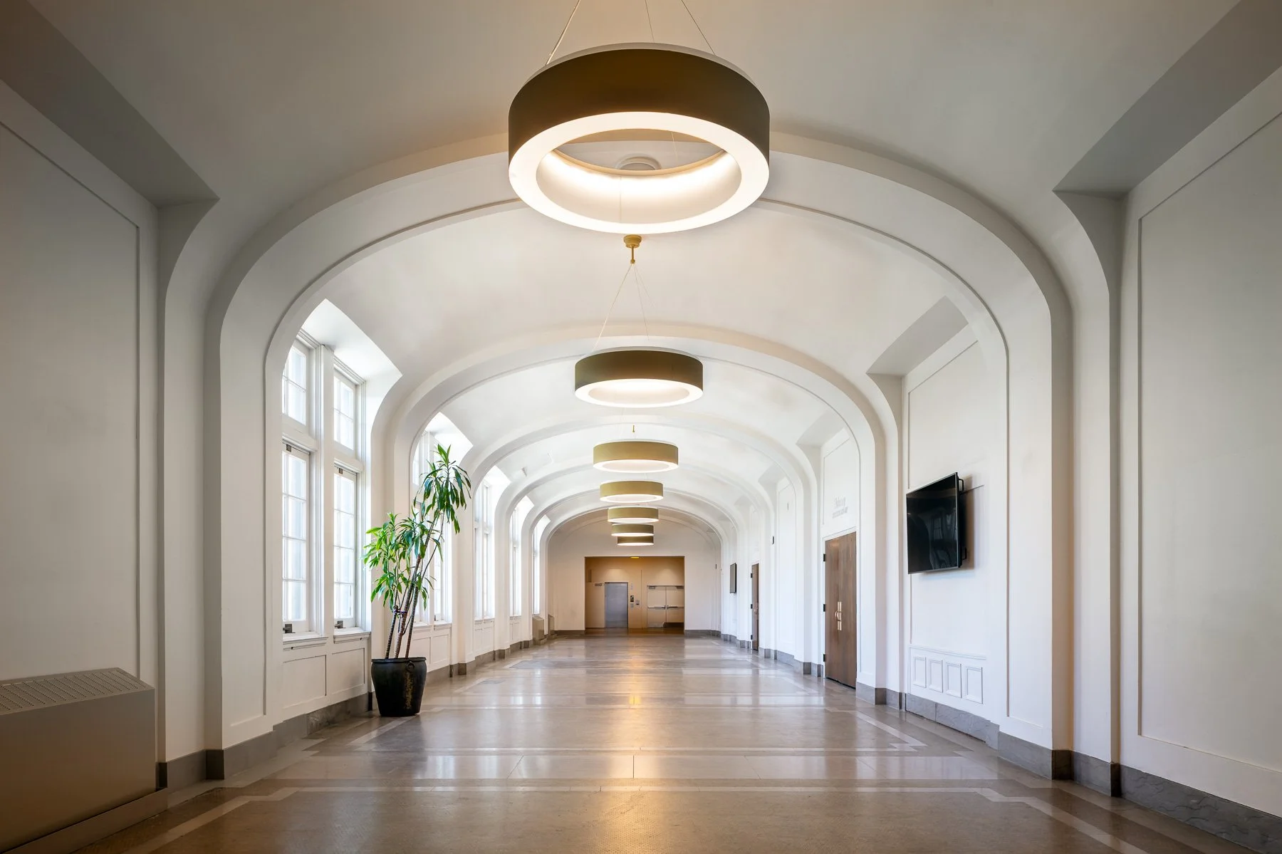 Empty hallway with arched ceiling, large windows, modern circular ceiling lights, potted plant, and a TV mounted on the wall.