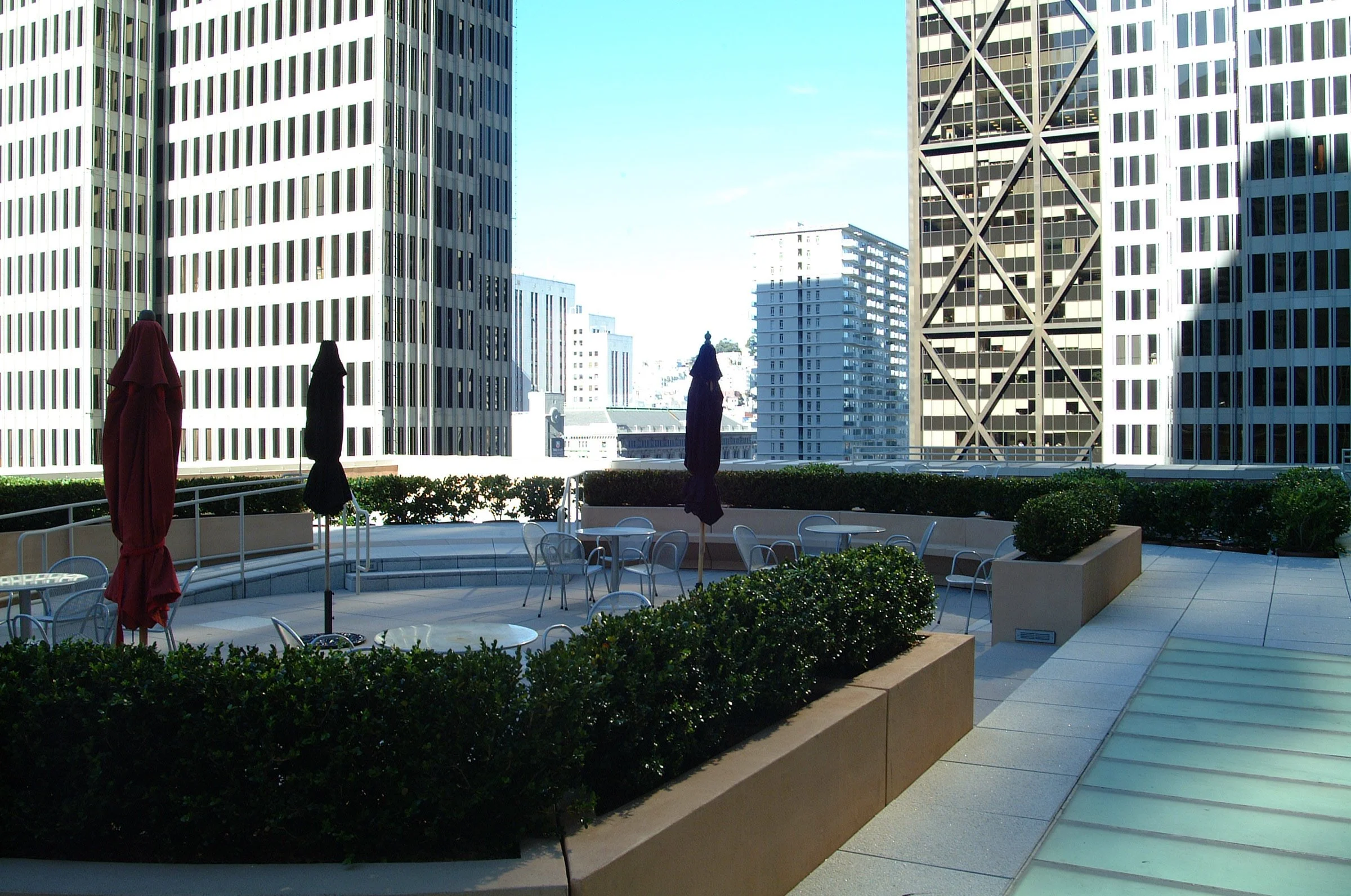 Empty outdoor patio on a high-rise building terrace with closed umbrellas, round tables, and chairs, surrounded by shrubs and tall city buildings.