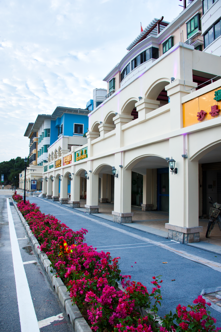 Colorful multi-story buildings with arched walkways, signs in Chinese, and pink flowers along the street