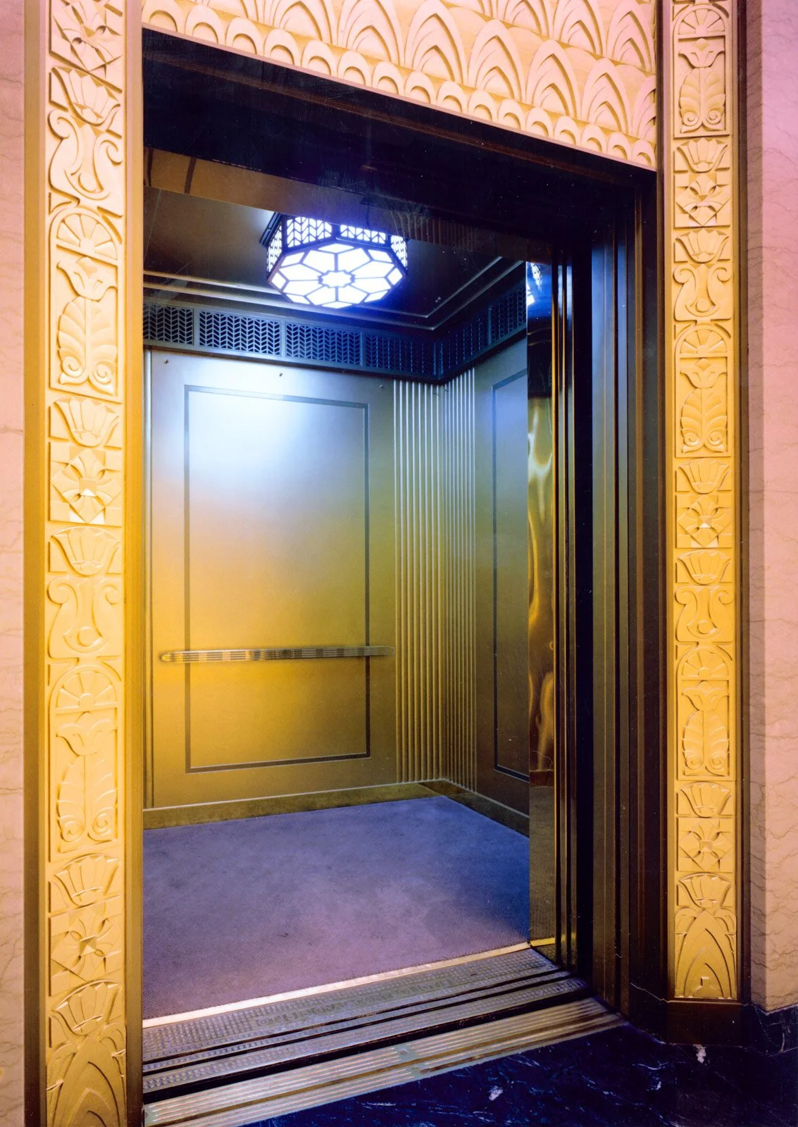 Elevator interior with gold-colored walls, a metal handrail, a patterned ceiling light, and a dark decorative ceiling grille.