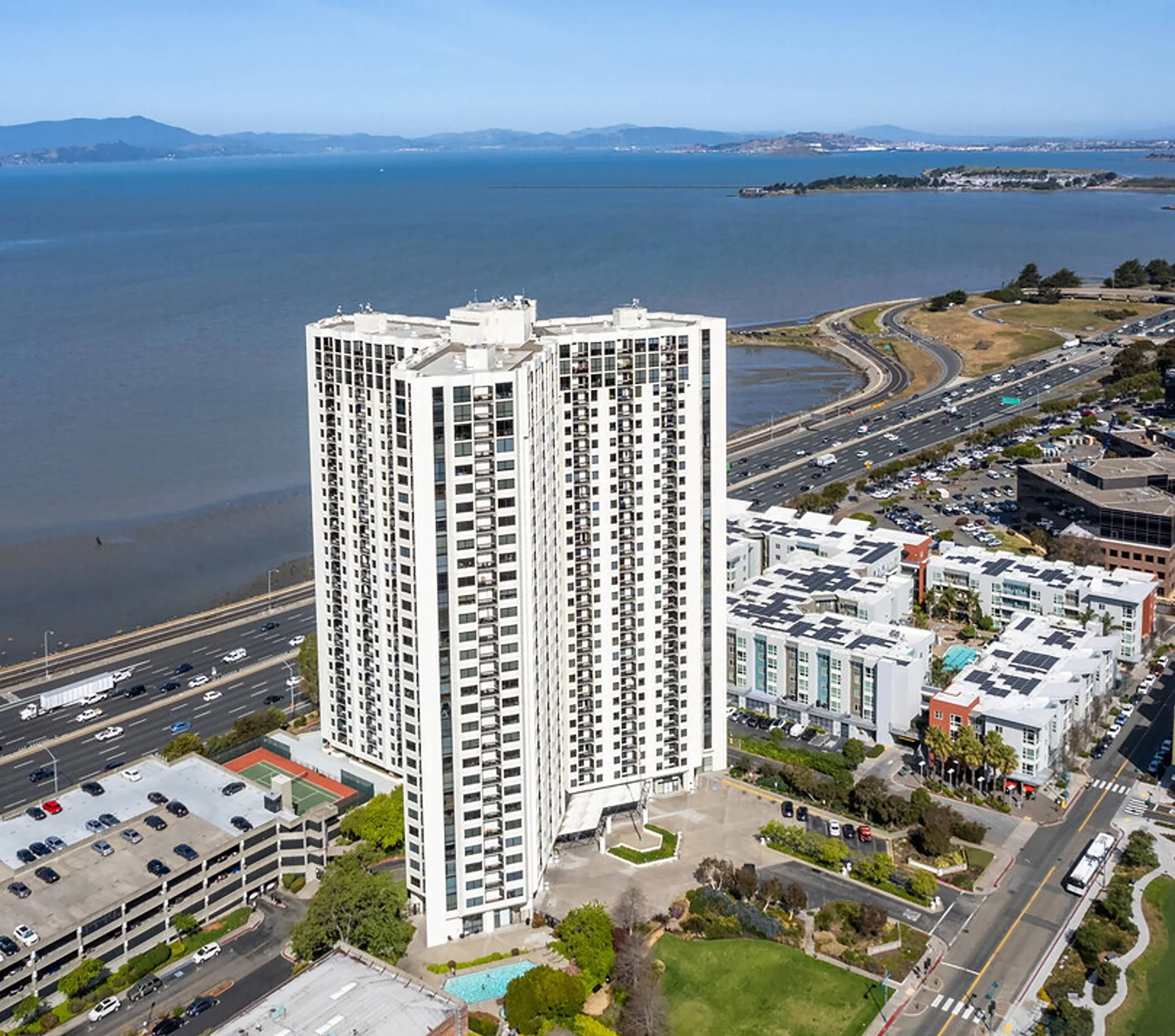 A tall white residential high-rise building facing a bay with mountains in the background. Nearby roads, parking lots, and smaller buildings are visible.