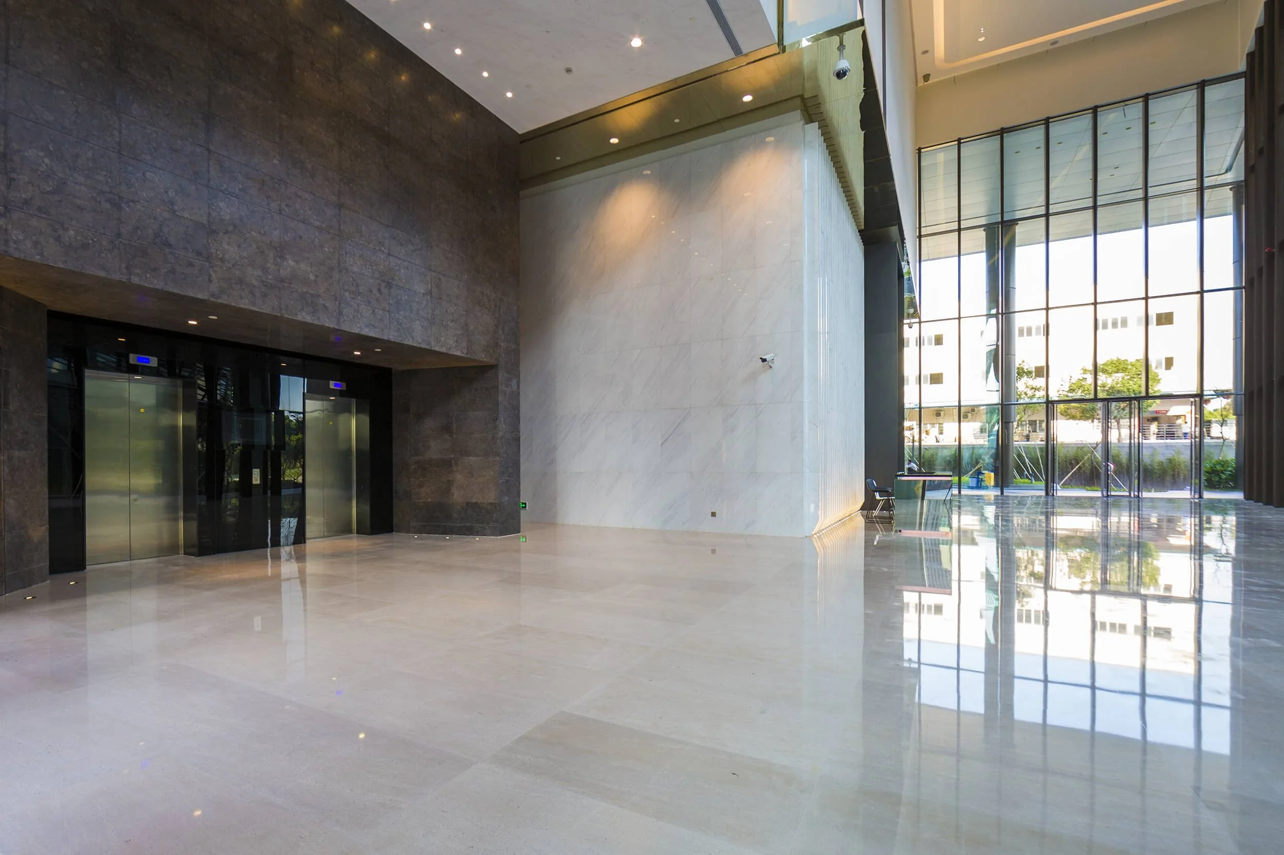 Empty modern lobby with large glass windows, shiny tiled floor, dark stone walls, and elevator doors on the left.