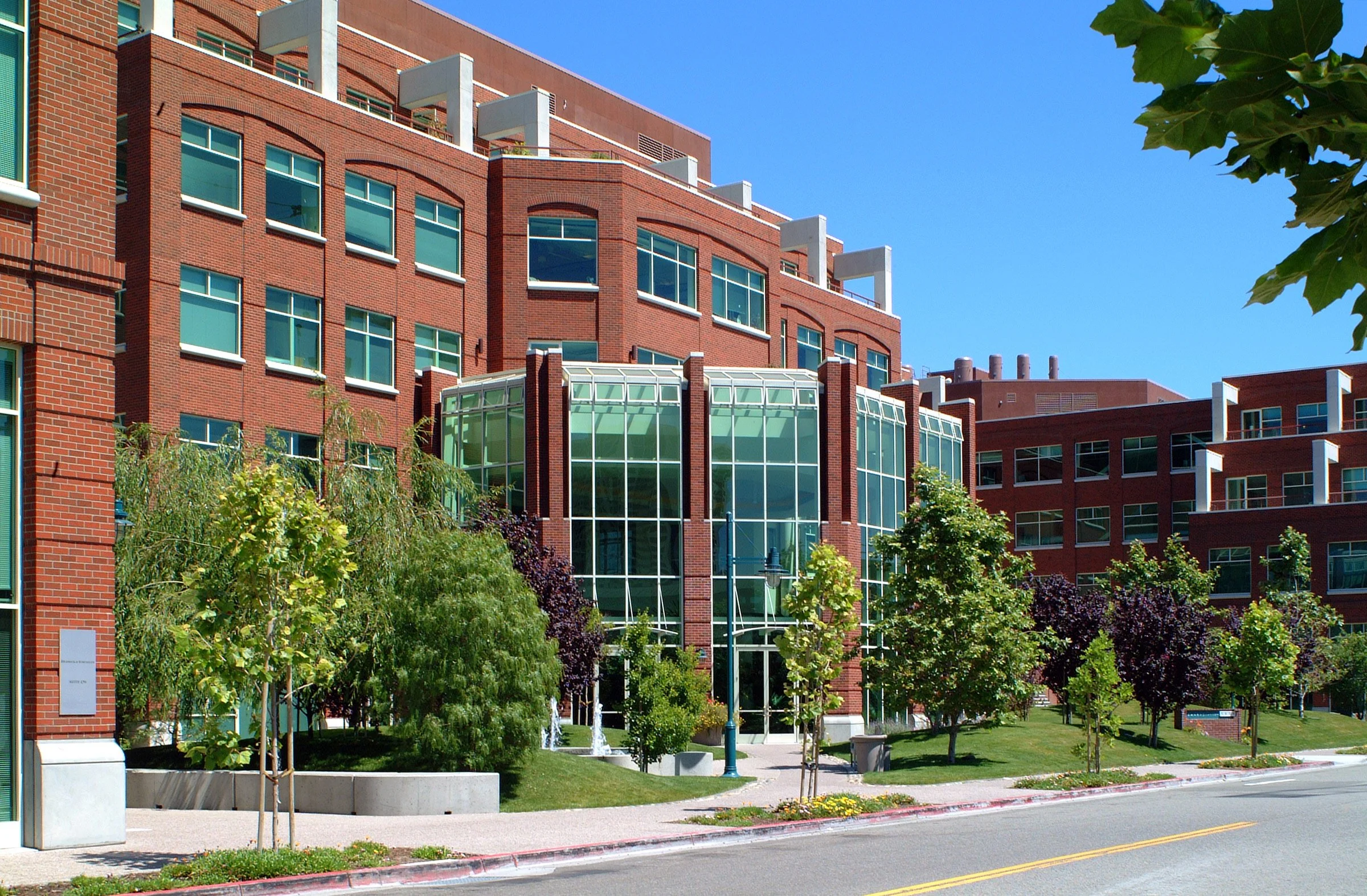 A modern multi-story building with large glass windows and red brick exterior, surrounded by trees and a well-maintained sidewalk.