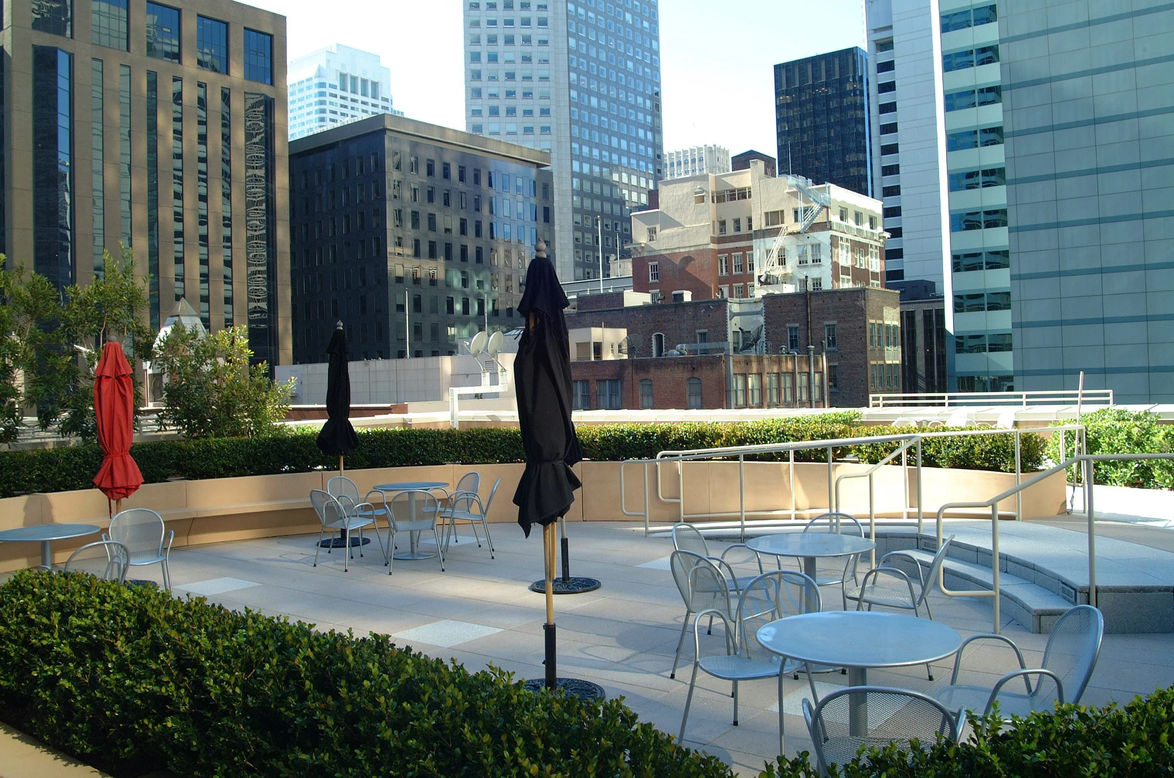 Empty rooftop outdoor seating area with tables, chairs, and umbrellas, surrounded by urban high-rise buildings and greenery.