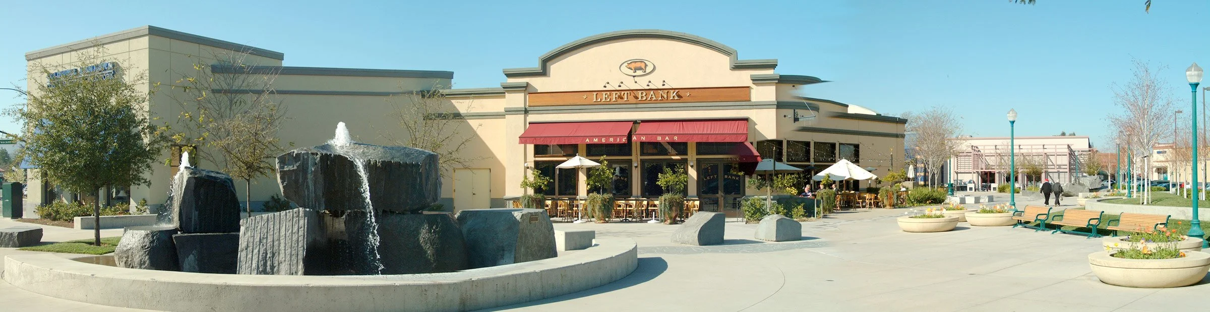 Outdoor shopping area with a water fountain, benches, trees, and a restaurant named 'Left Bank' with outdoor seating and umbrellas.