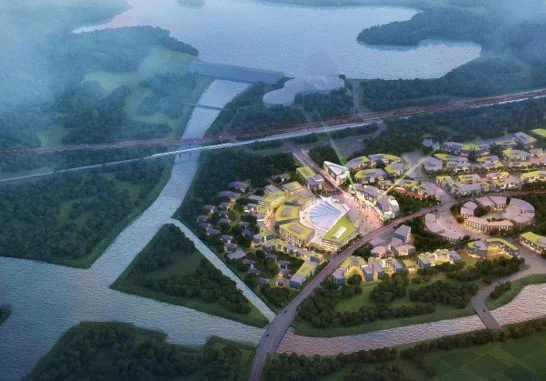 Aerial view of a cityscape with a large stadium, surrounding buildings, and roads, with a river and green spaces nearby.