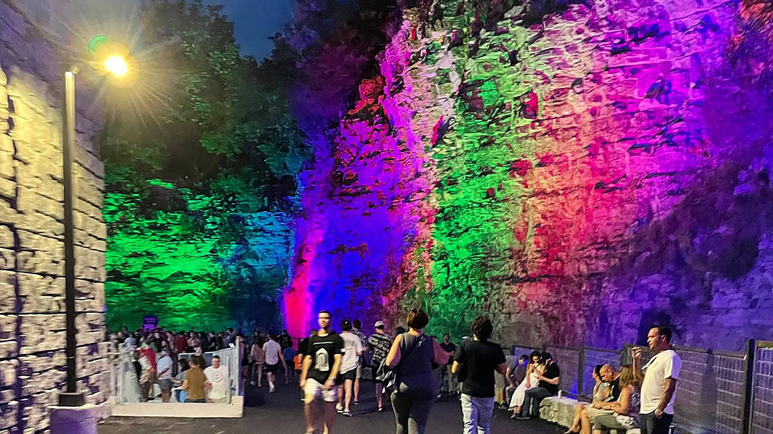 People walking and sitting near a large rock wall decorated with multicolored lighting at night.