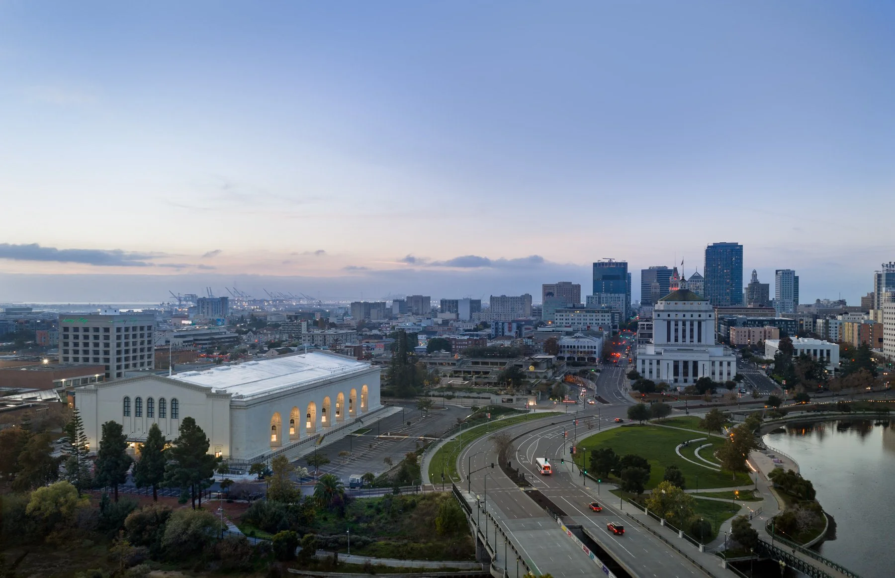 A cityscape at dusk featuring a large white government building, surrounding parking lots, roads with cars, a park with a pond, and a skyline of tall office buildings in the background.