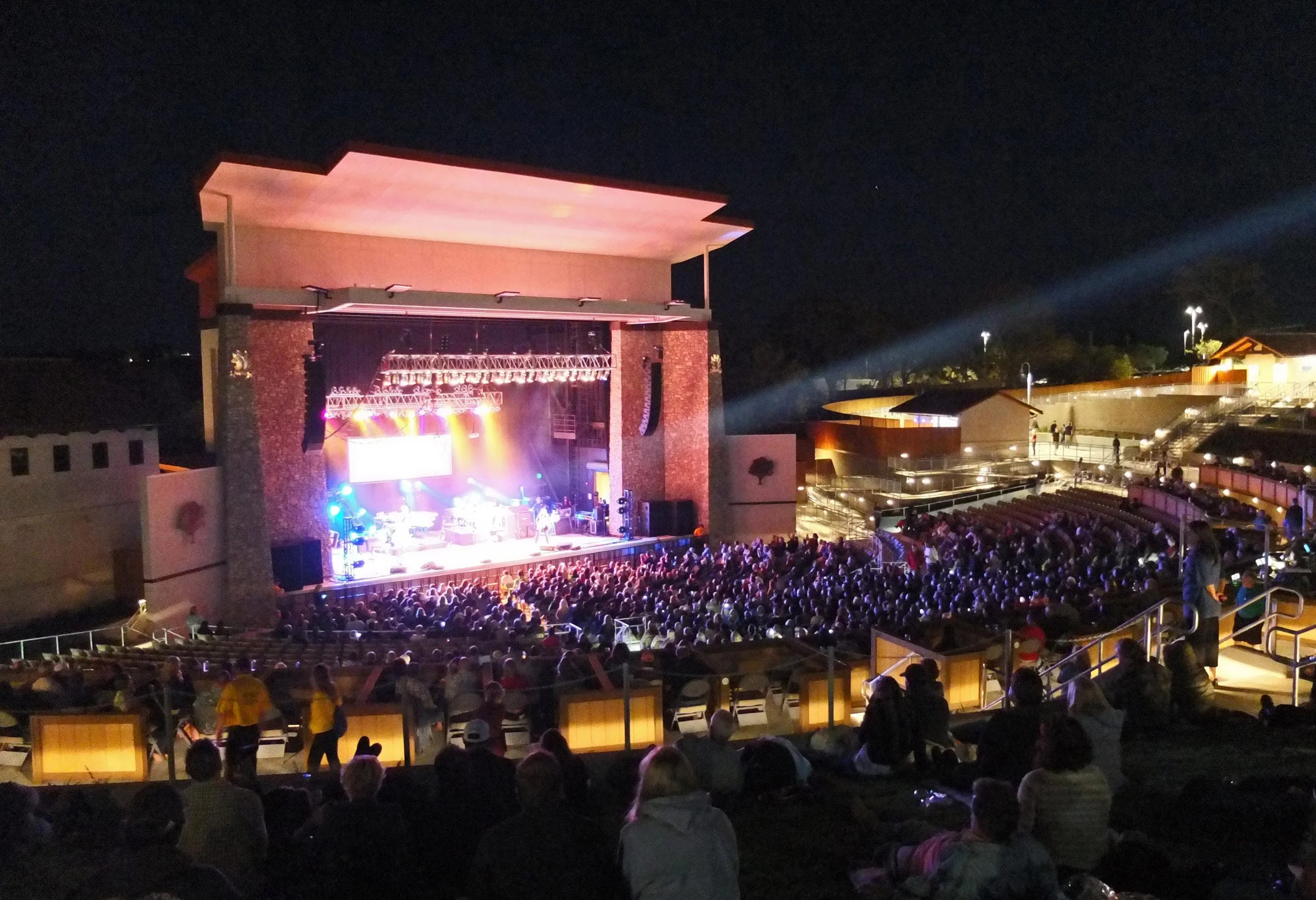 An outdoor amphitheater at night with a live concert on stage and a large audience seated in front, illuminated by stage lights.