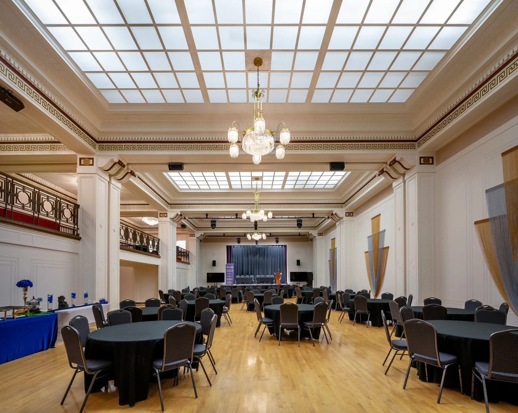 Elegant banquet hall with round tables covered in black cloth, black chairs, chandeliers, and a stage at the front, decorated with blue curtains.