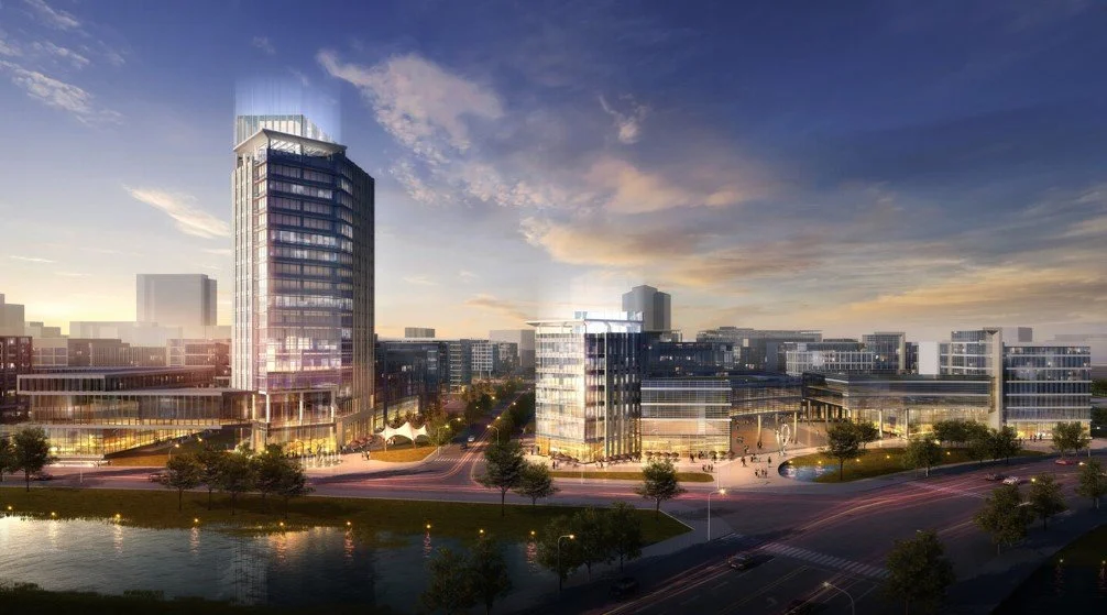 A city landscape at dusk featuring modern glass buildings, a river in the foreground, and a partly cloudy sky with the sun setting.