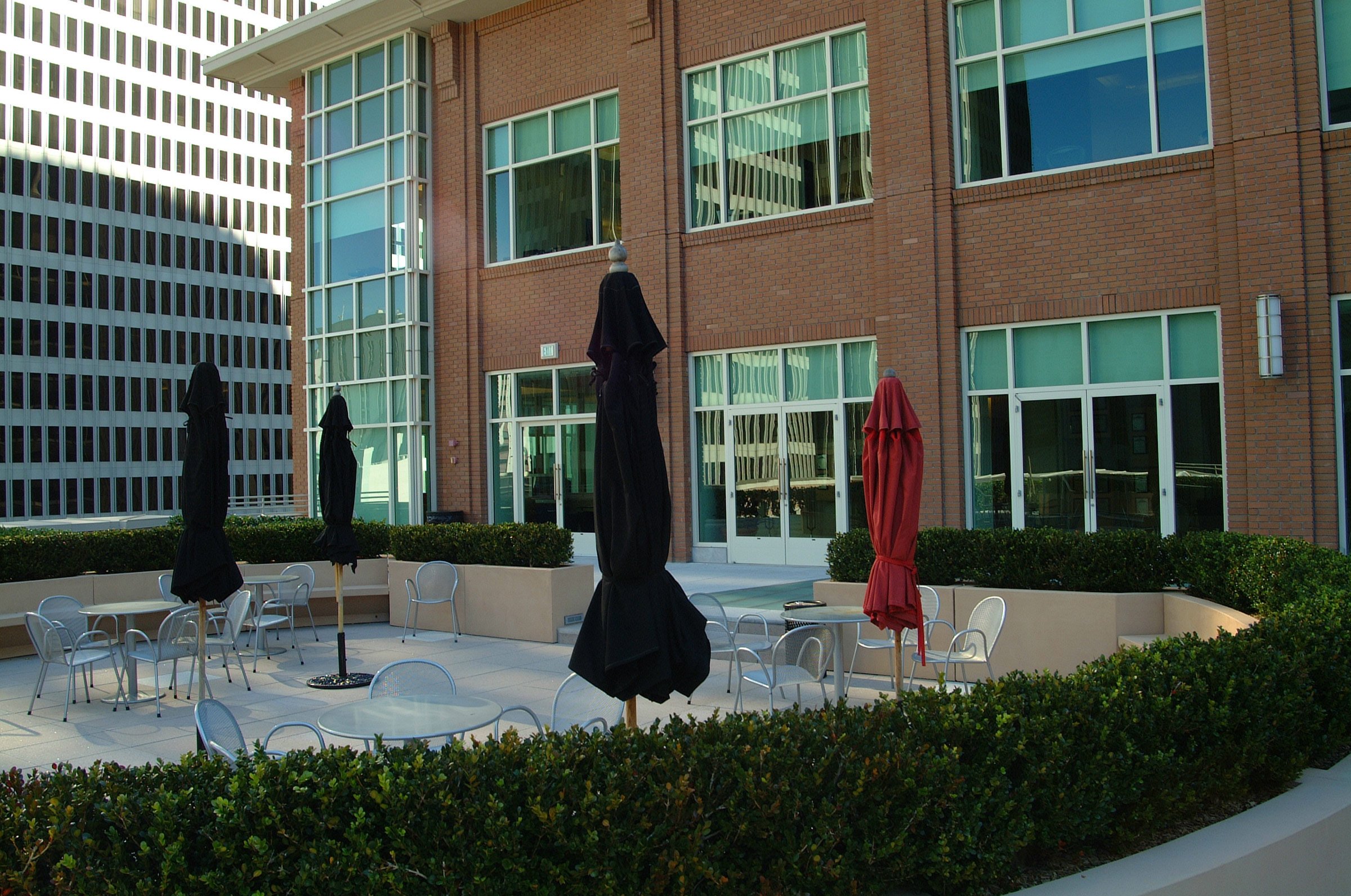 Outdoor rooftop seating area with round tables, chairs, and closed umbrellas in front of a modern brick building with large windows.