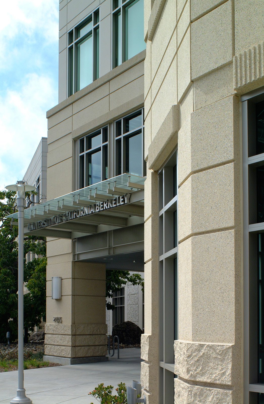 Exterior of the University of California Berkeley building with beige stone walls, large windows, and a sign reading 'UNIVERSITY OF CALIFORNIA BERKELEY' on a glass canopy.