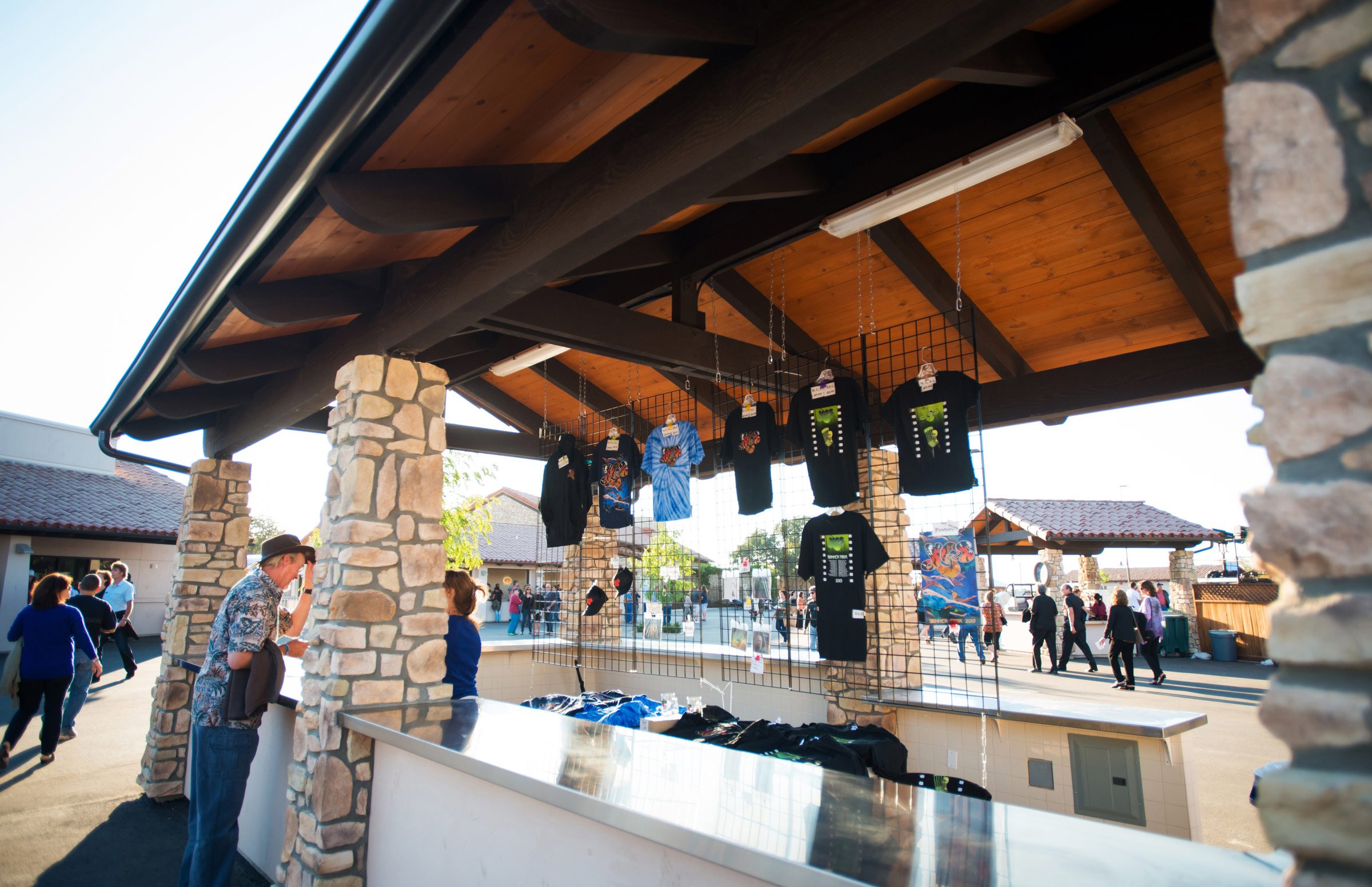 Mercado de pulgas con camisetas y otros artículos en exhibición, personas caminando y vendiendo, estructuras de piedra y madera bajo un techo de madera